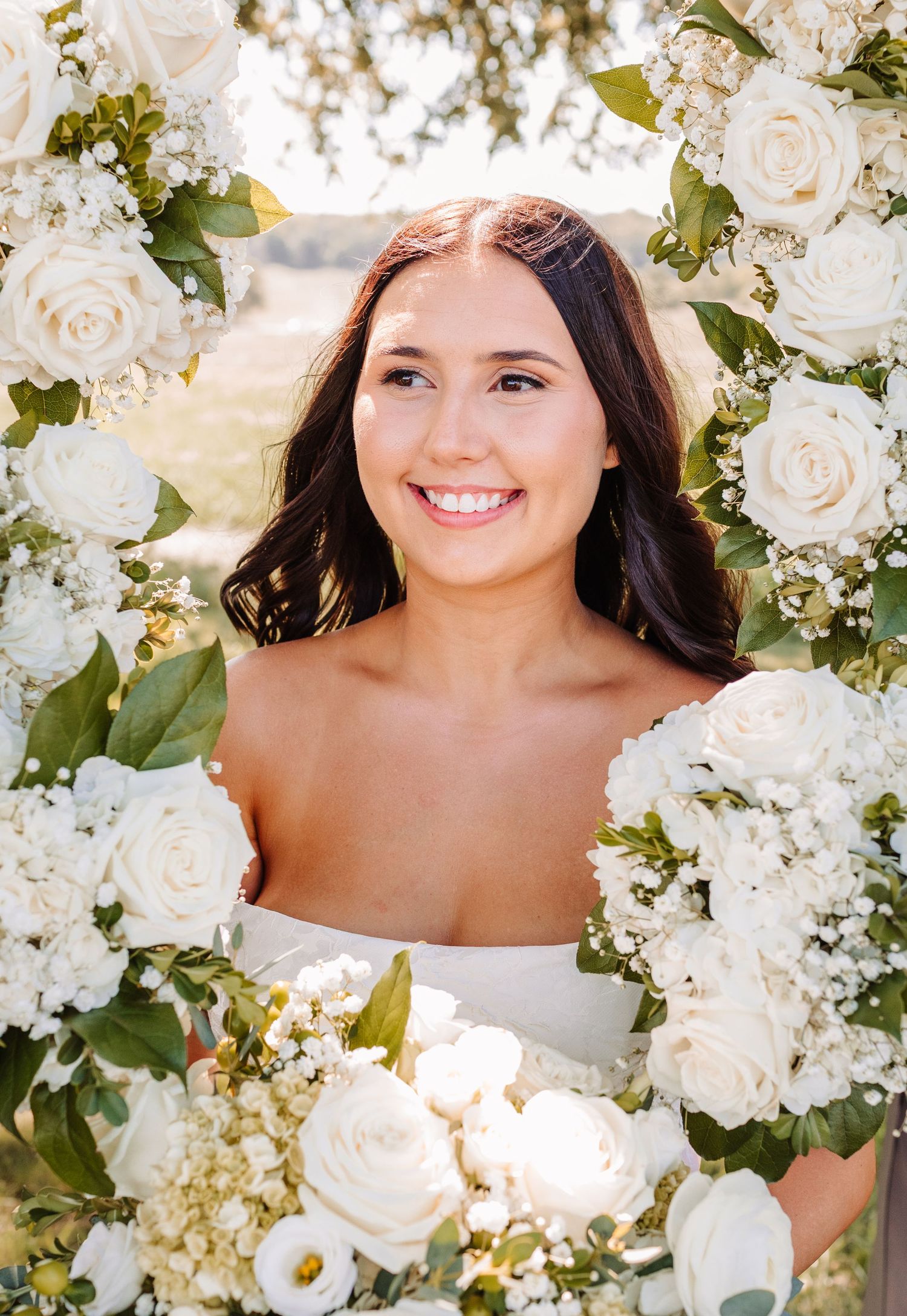 Smiling bride holding white roses bouquet framed by floral arch with white roses and greenery on wedding day.