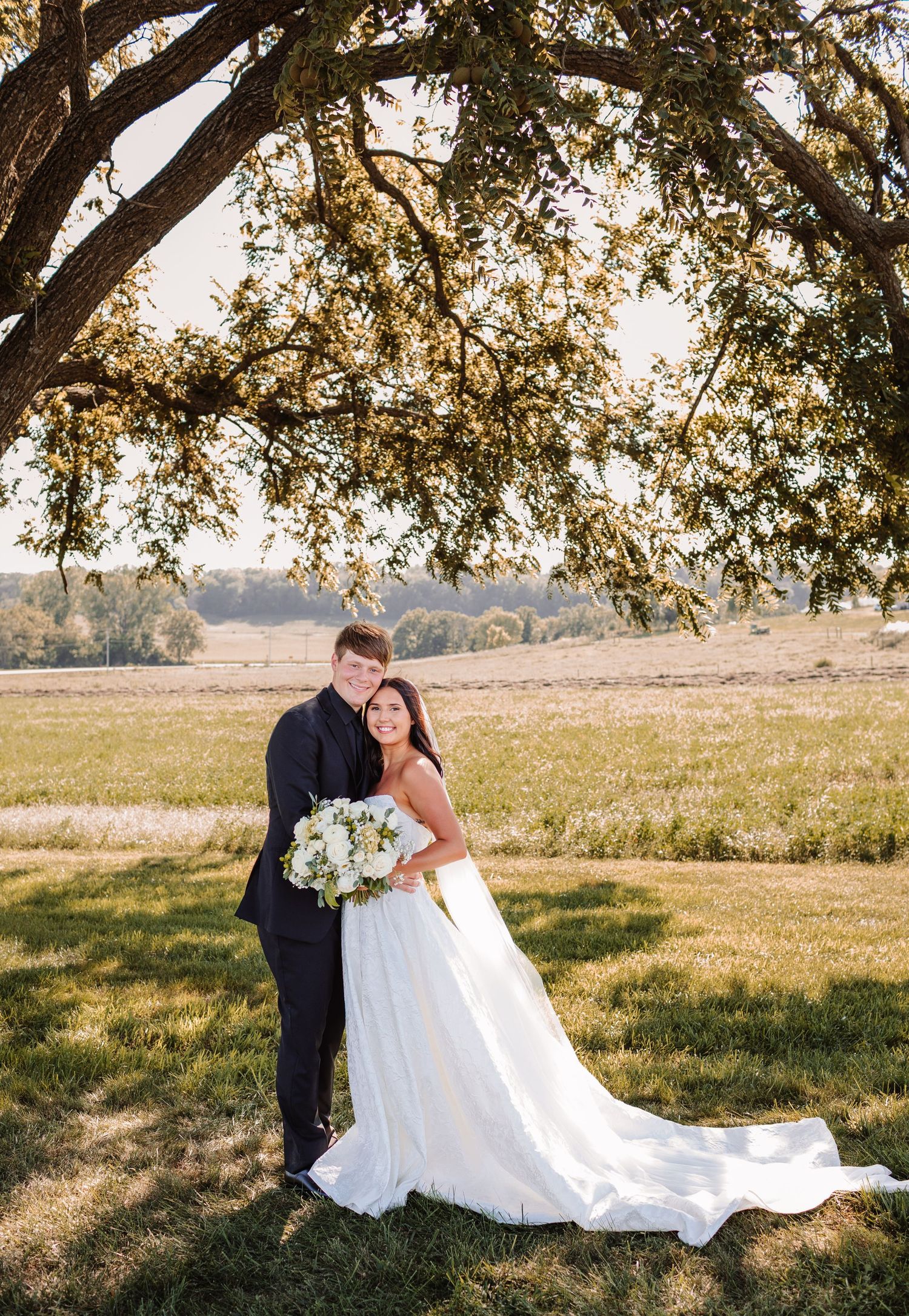Bride and groom pose under a large oak tree in a sunlit field, bride holding a white bouquet in a flowing gown.