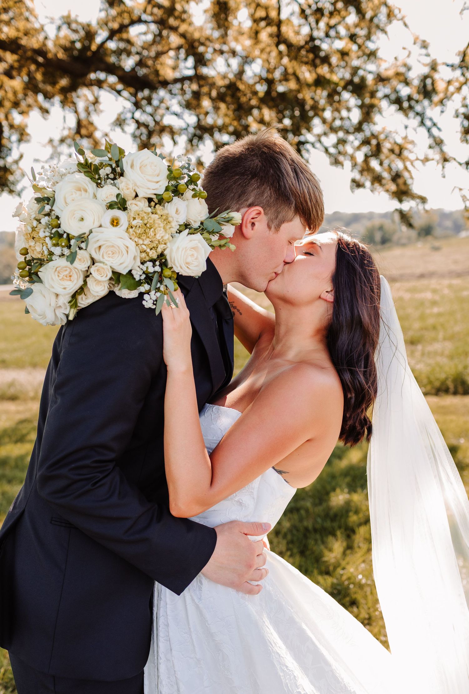 A bride and groom share a romantic kiss outdoors, with a white rose bouquet and flowing veil under a sunlit tree.