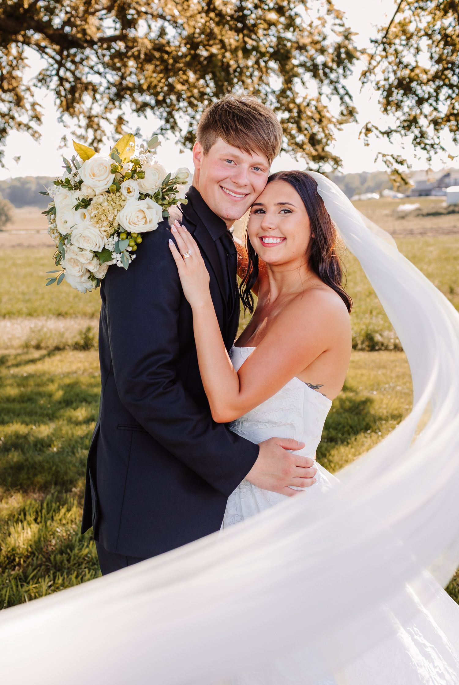 Smiling bride and groom embrace outdoors, her veil flowing in the wind, holding white bouquet under a tree at golden hour.