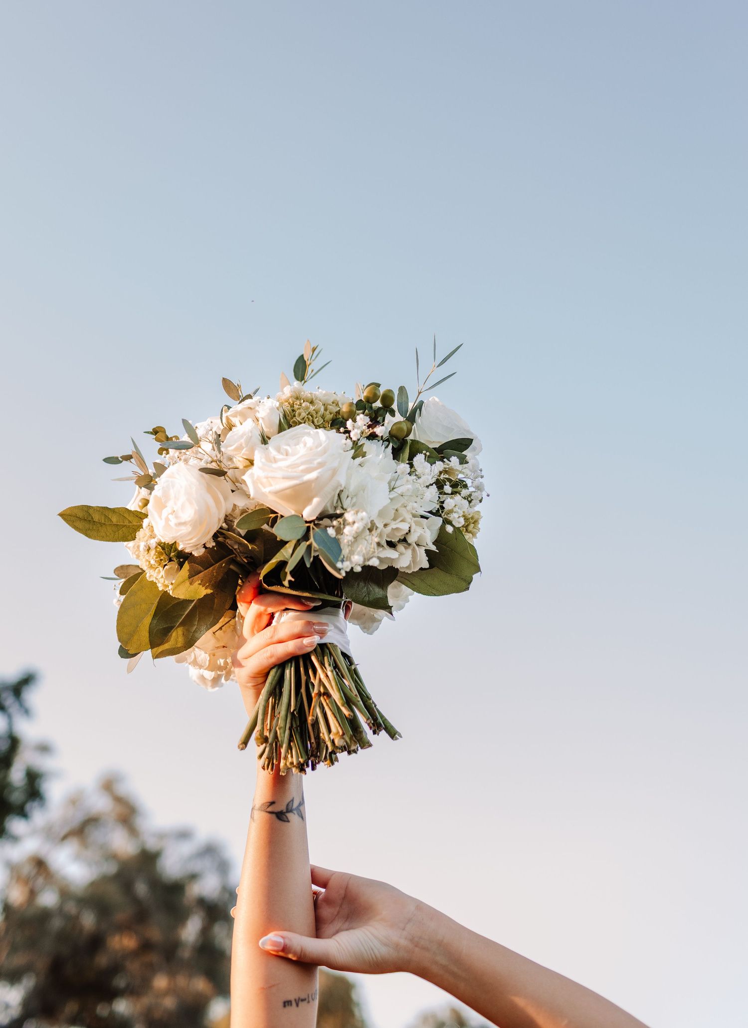 A hand holding a bridal bouquet of white roses and greenery against a clear blue sky.