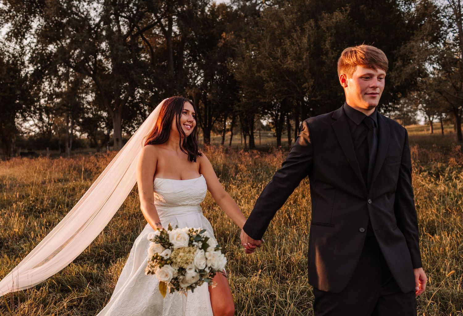 Bride and groom holding hands walking through golden field at sunset, bride wearing white gown with veil holding bouquet.