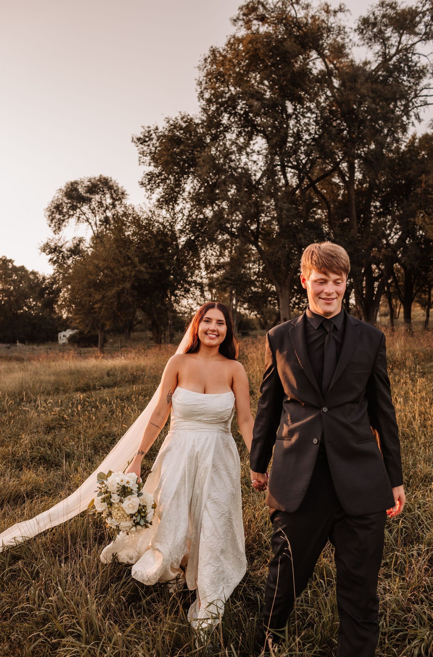 A smiling bride in a white gown and groom in a black suit hold hands in a golden field at sunset.