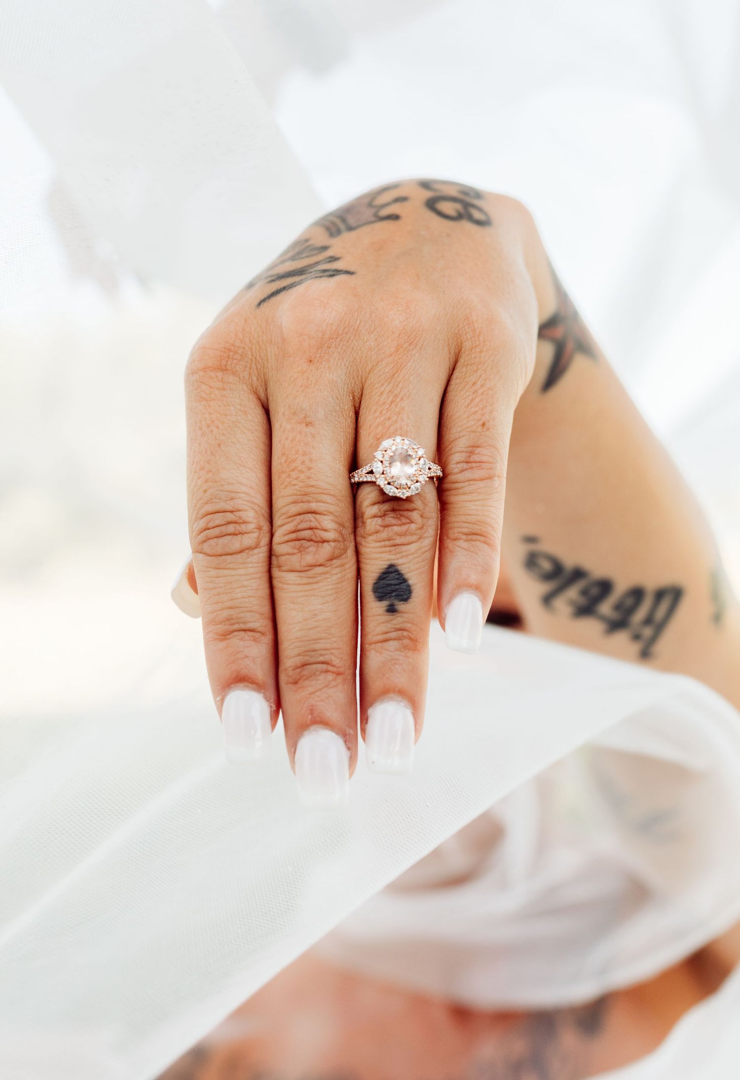 Tattooed bride showcasing a round halo engagement ring with white manicured nails on wedding day.