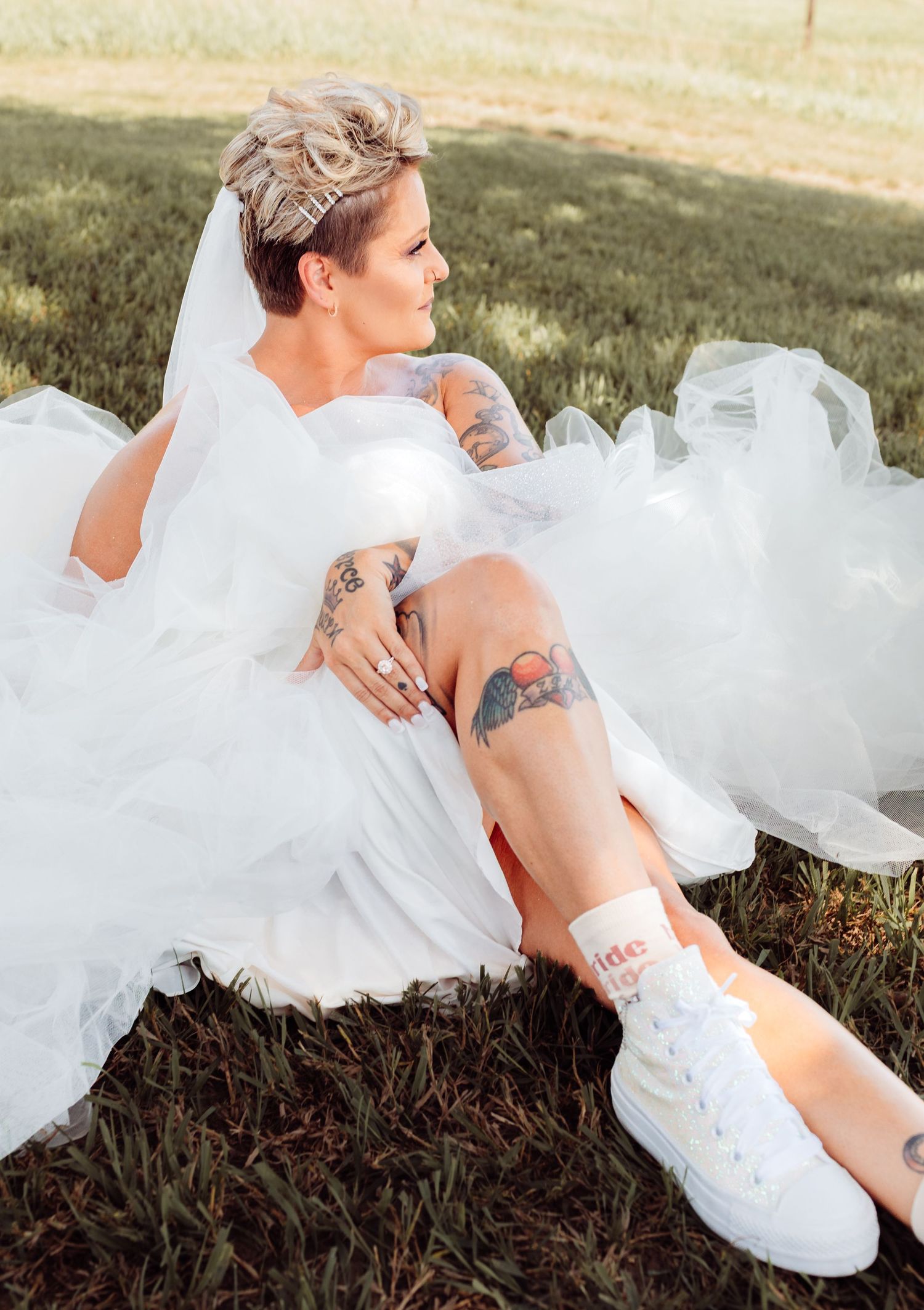 Tattooed bride in white wedding gown and sneakers sitting on grass wearing a veil outdoors.
