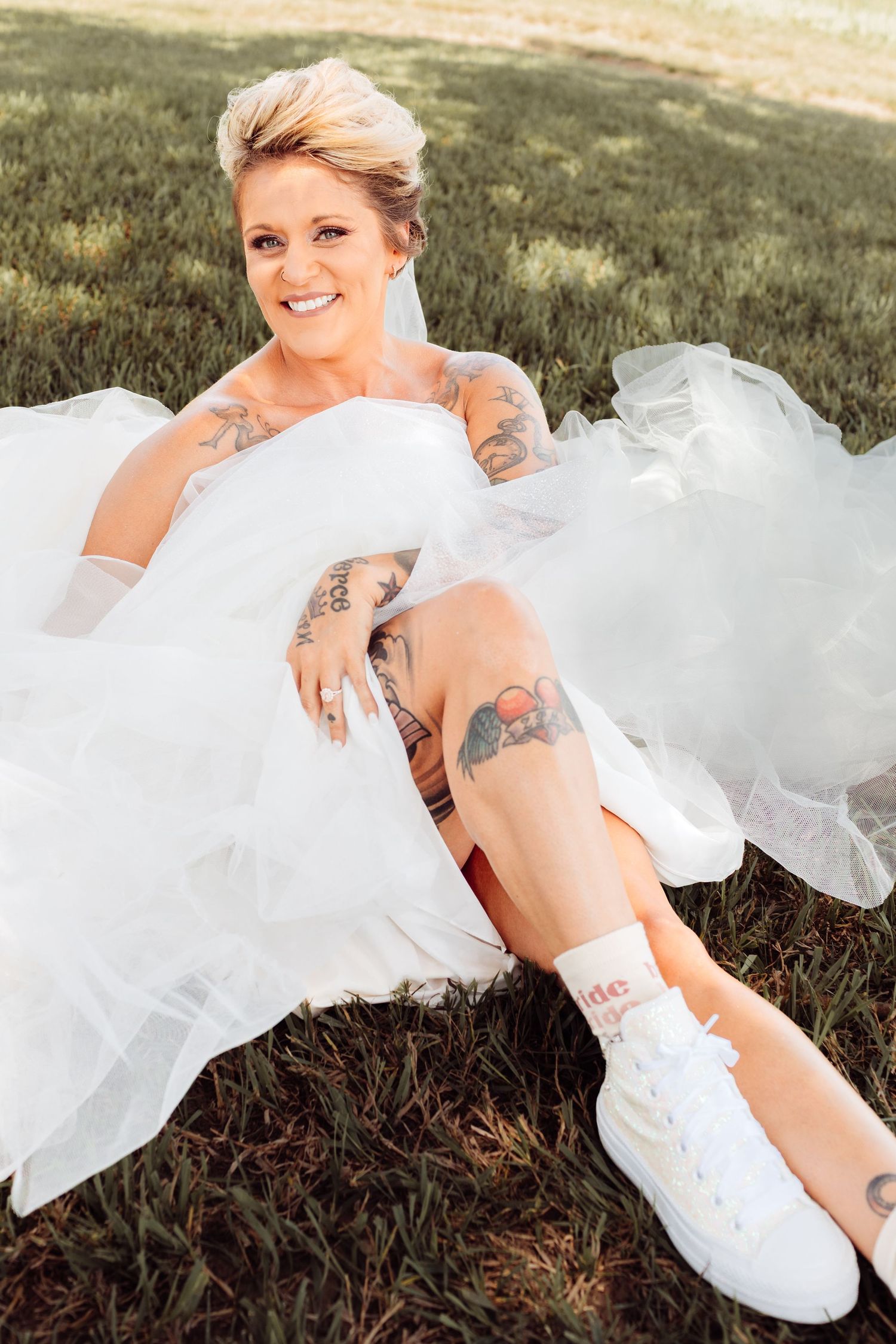 Smiling tattooed bride in white wedding dress and white sneakers sitting on grass outdoors.