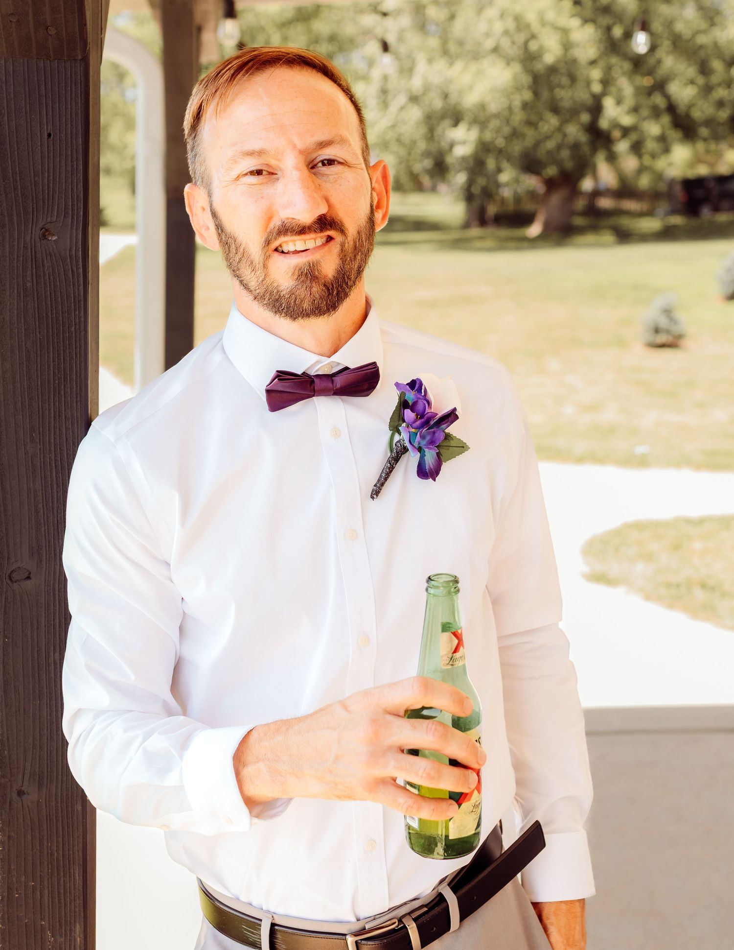 Smiling groom in white shirt with purple bow tie and boutonniere holding a green bottle.