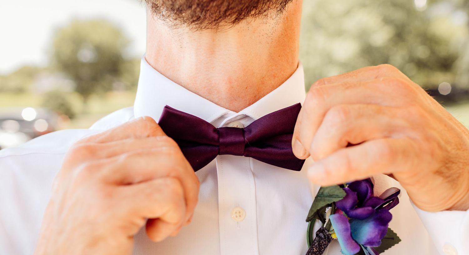 Groom adjusting purple bow tie with floral boutonniere on white dress shirt.