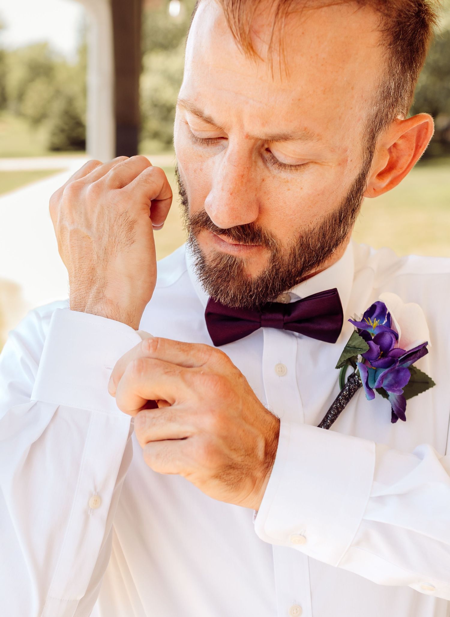 Groom in white shirt fastening cuff links with purple bow tie and blue boutonniere.