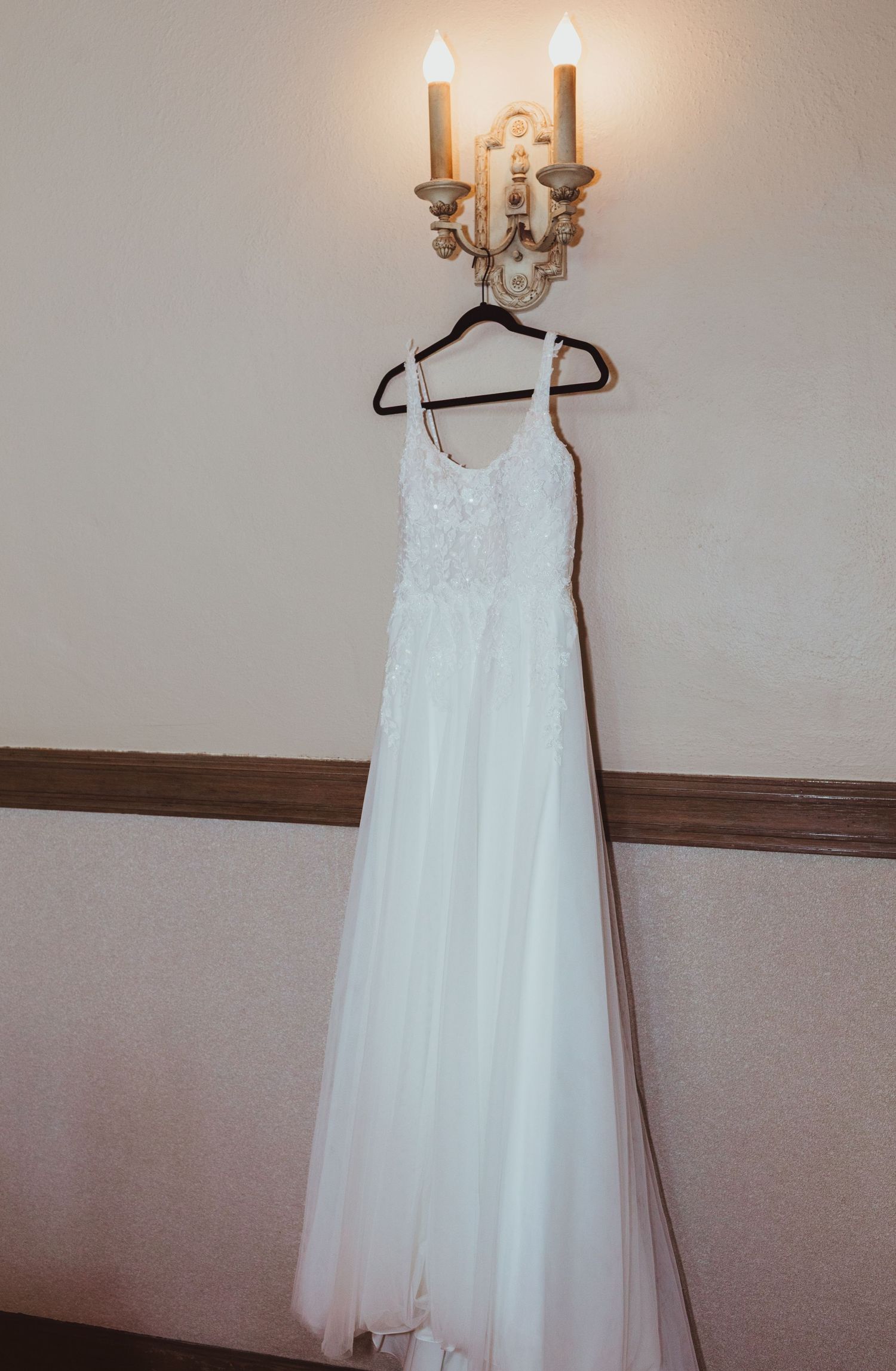 Elegant white lace bridal gown displayed on a hanger beneath a decorative candle wall sconce.