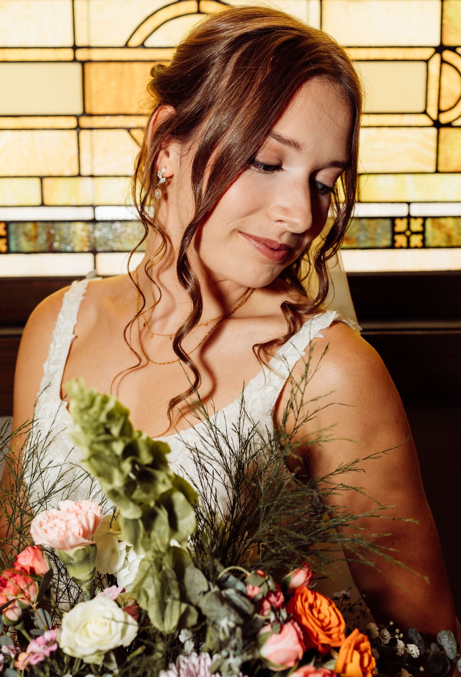 Beautiful bride with soft curled hair holding a colorful bouquet before a stained glass window.