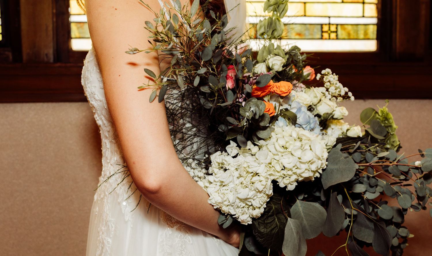 Bride in white lace gown holding a lush bouquet of white hydrangeas and orange roses.