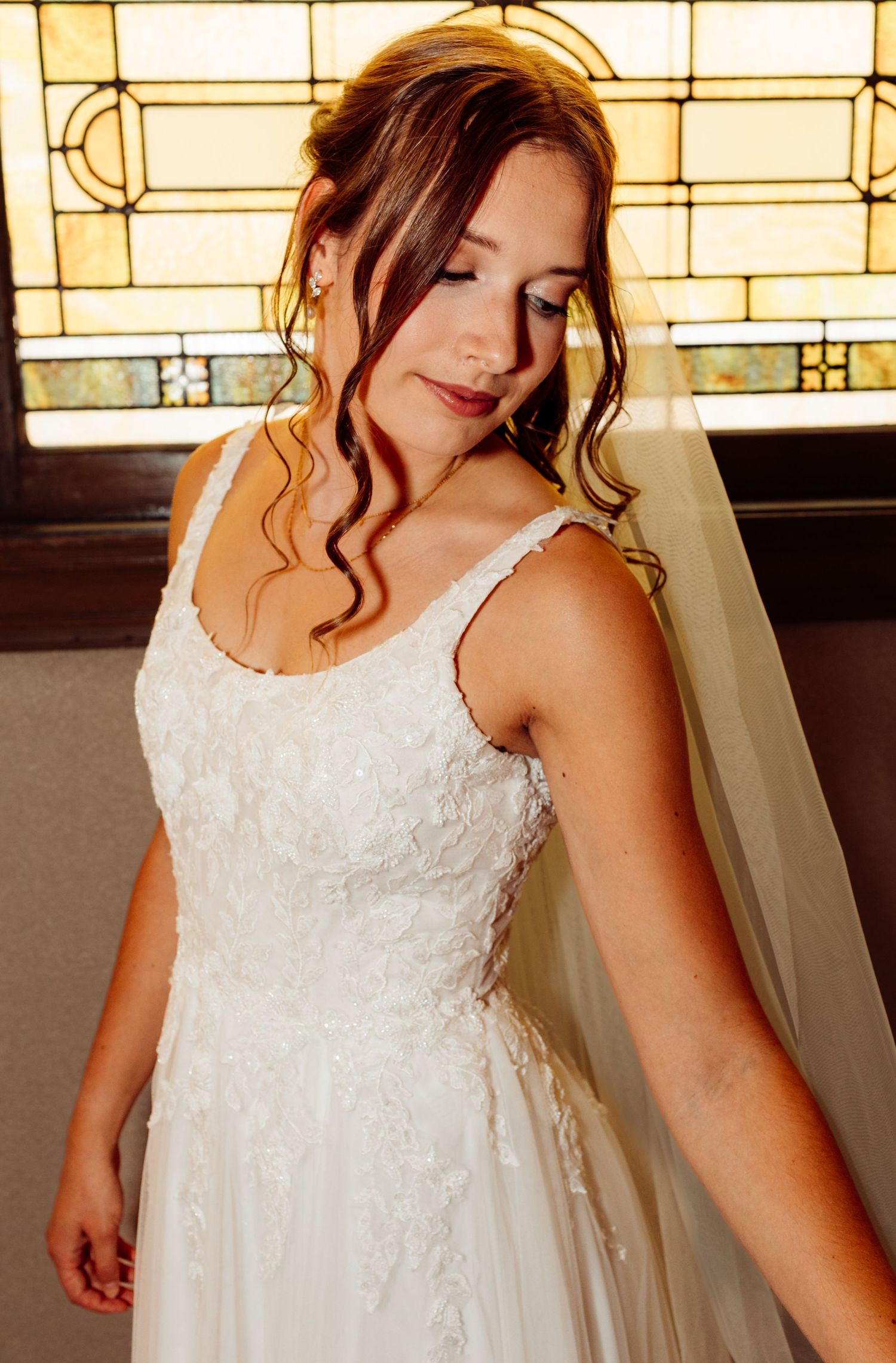 Elegant bride in a white lace gown with veil standing before a stained glass church window.