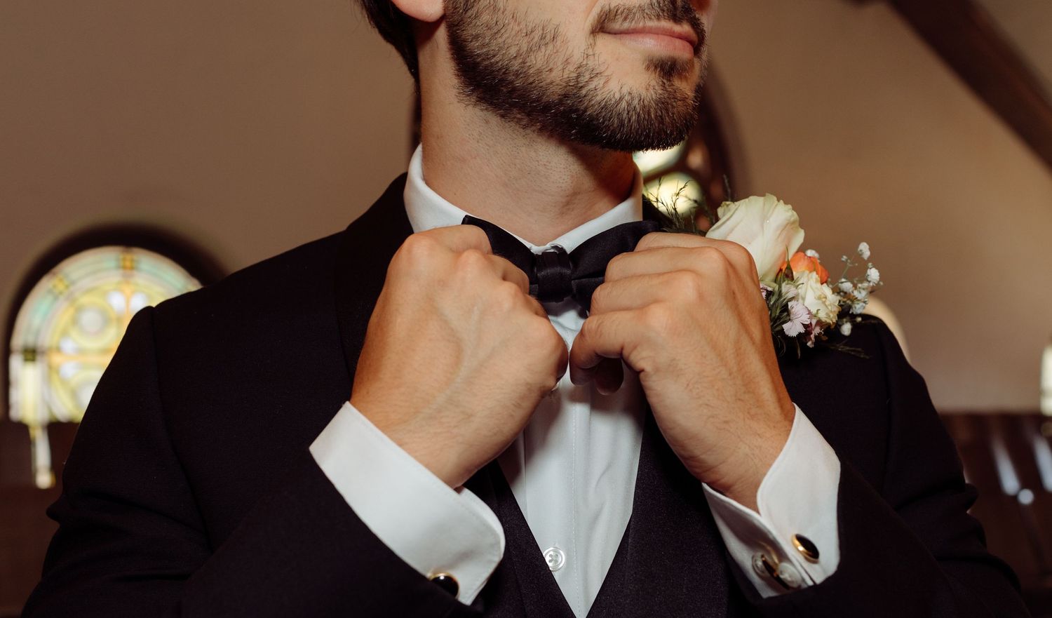 Groom in black tuxedo adjusting his bow tie with a floral boutonniere on his lapel.