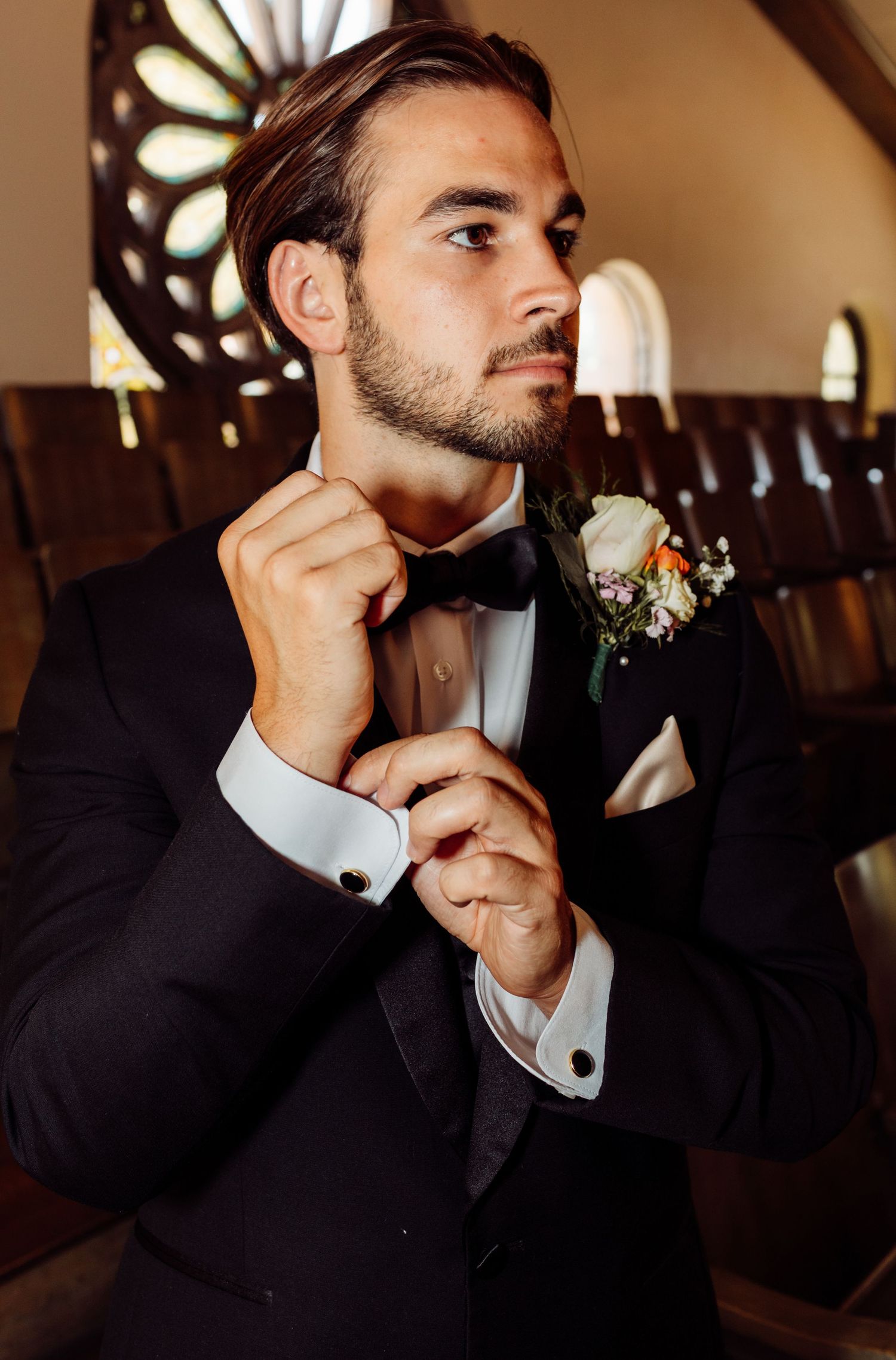 Groom in black tuxedo adjusting bow tie with boutonniere in church before wedding ceremony.