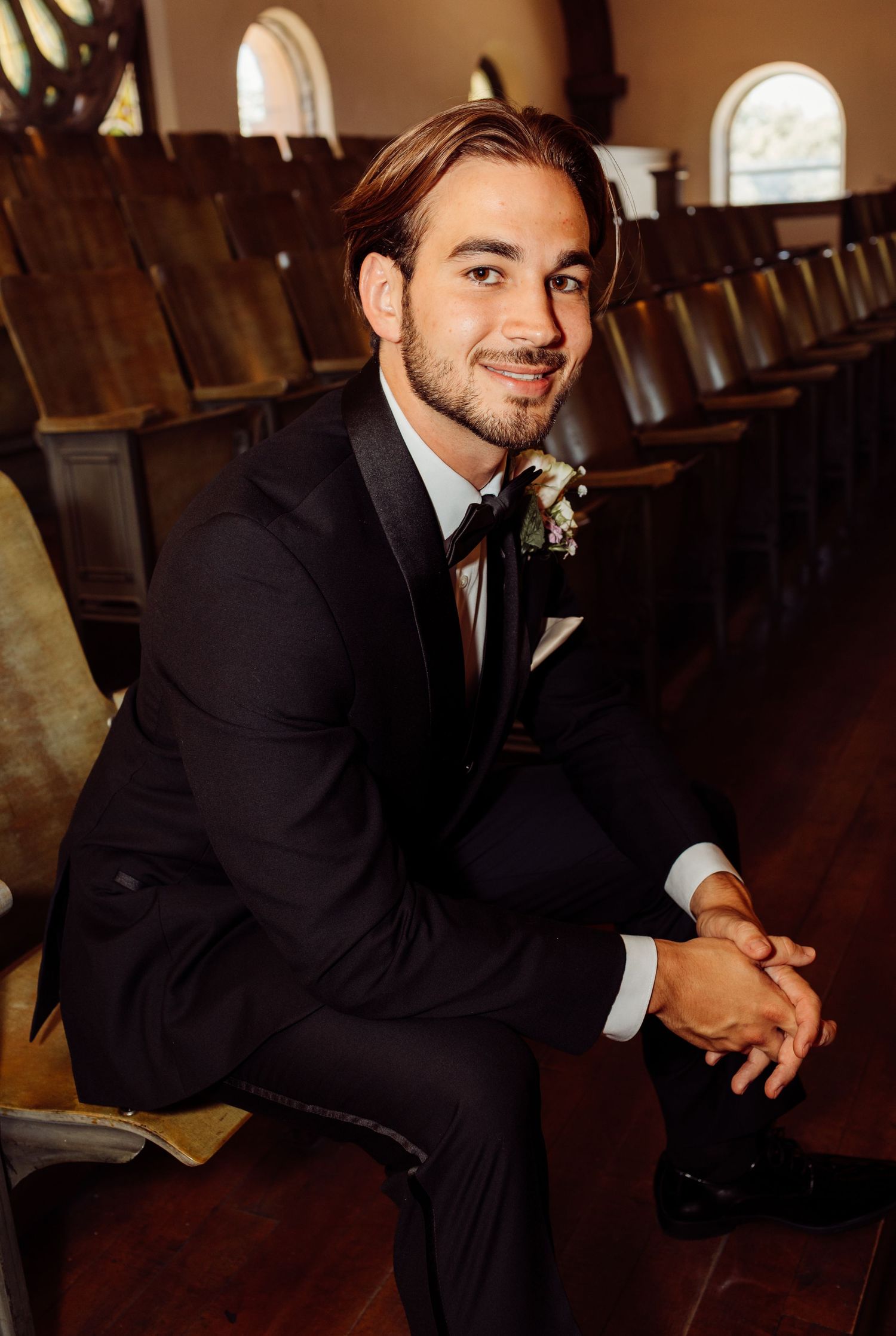 Handsome groom in a black tuxedo sitting in church pews with a floral boutonniere.