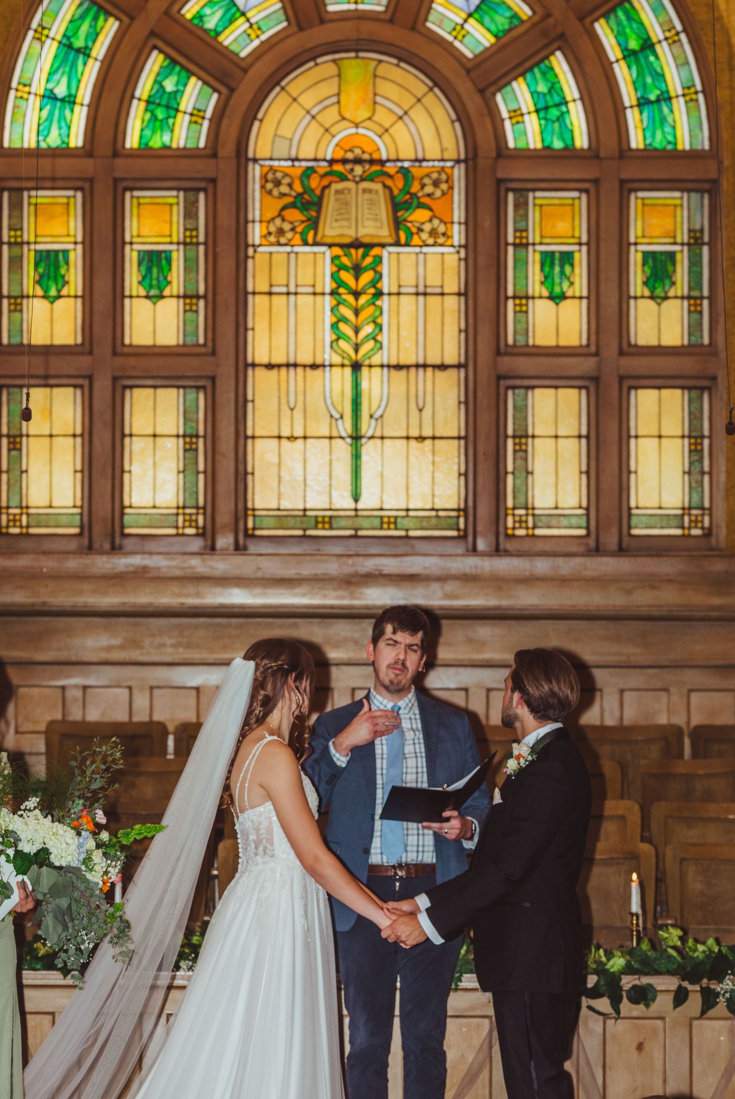 Bride and groom exchange vows before stunning stained glass window with officiant at church altar.