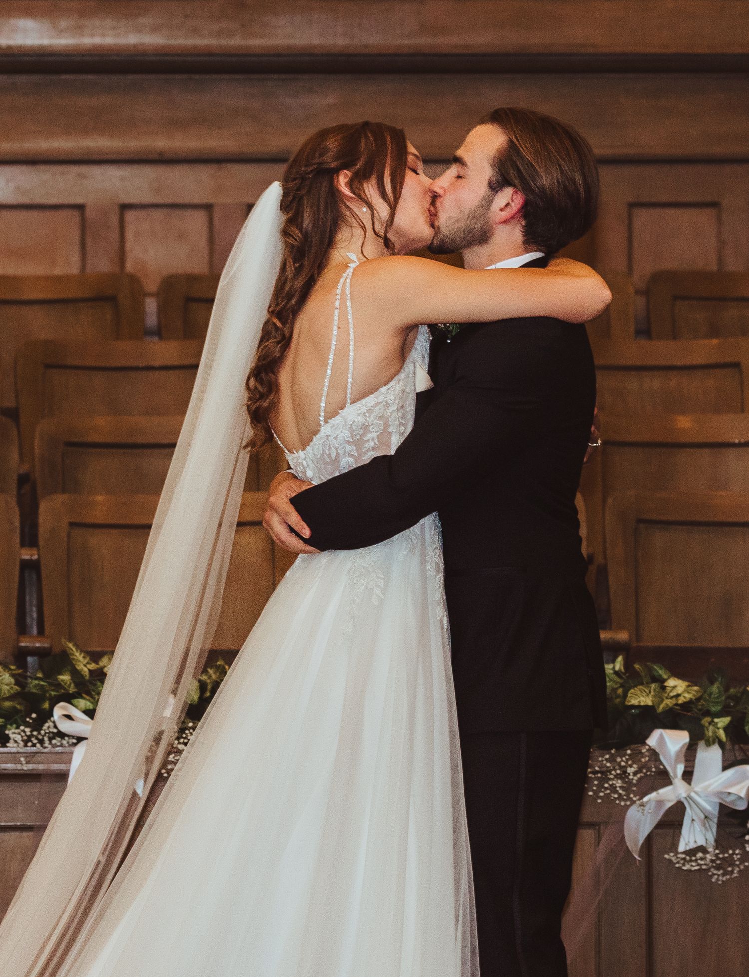 Bride and groom share first kiss in church ceremony, bride wearing lace wedding gown with veil.