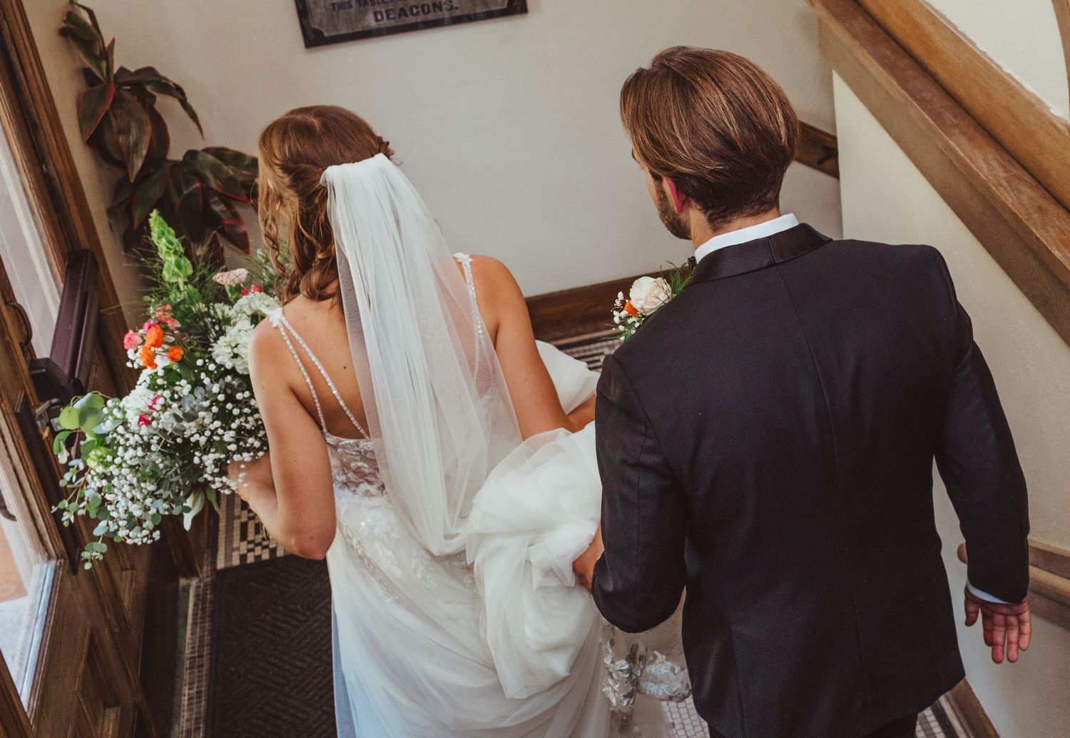 Bride and groom walk hand in hand up church stairs, bride holding colorful floral bouquet.