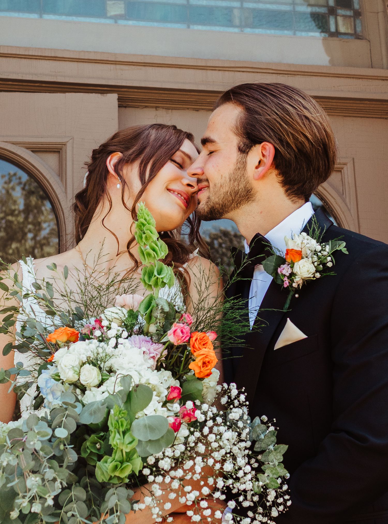 Newlyweds share romantic kiss surrounded by vibrant colorful wedding bouquet outside church.