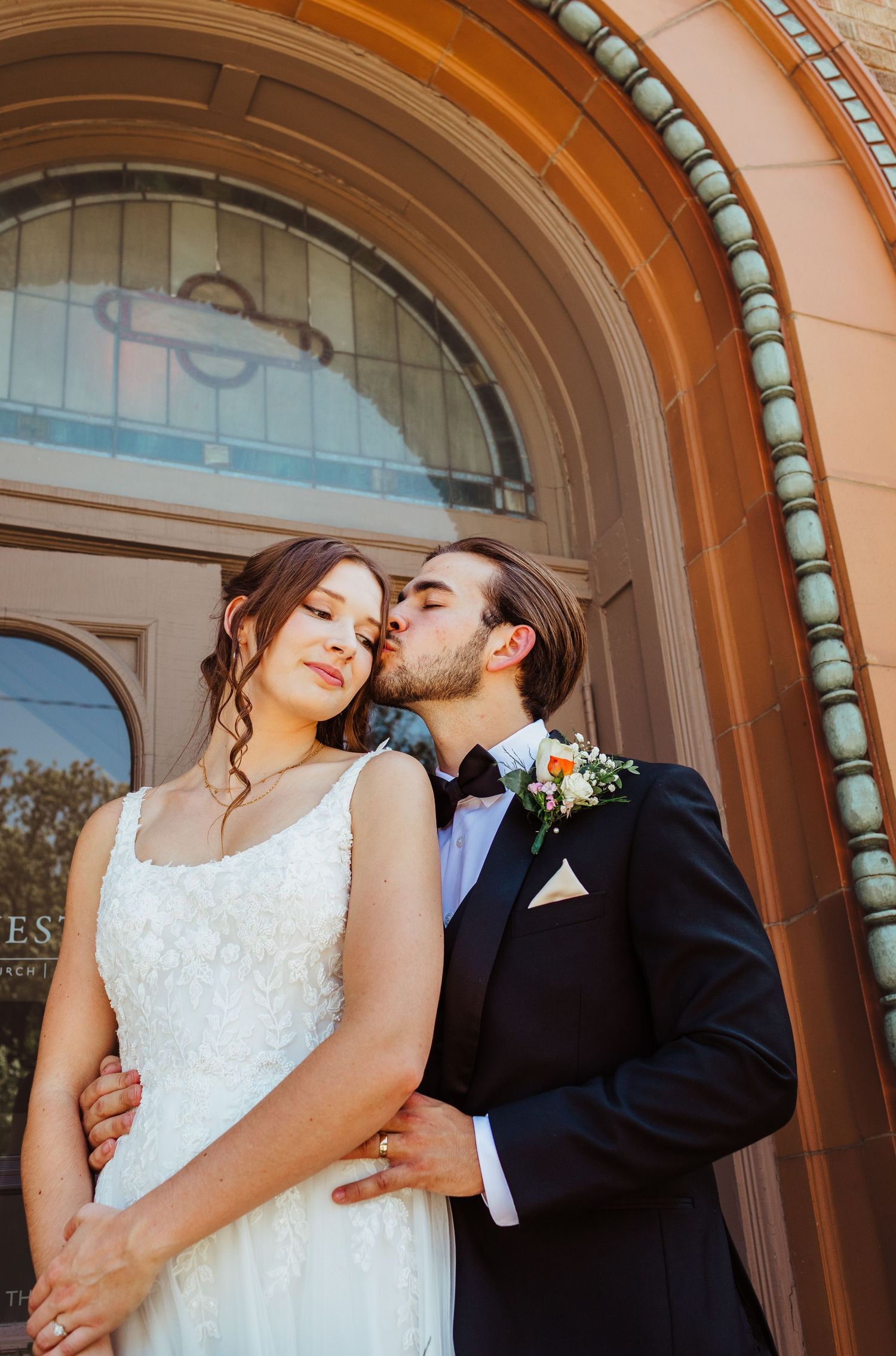 Groom in black tuxedo kisses bride in lace gown under ornate church archway entrance.