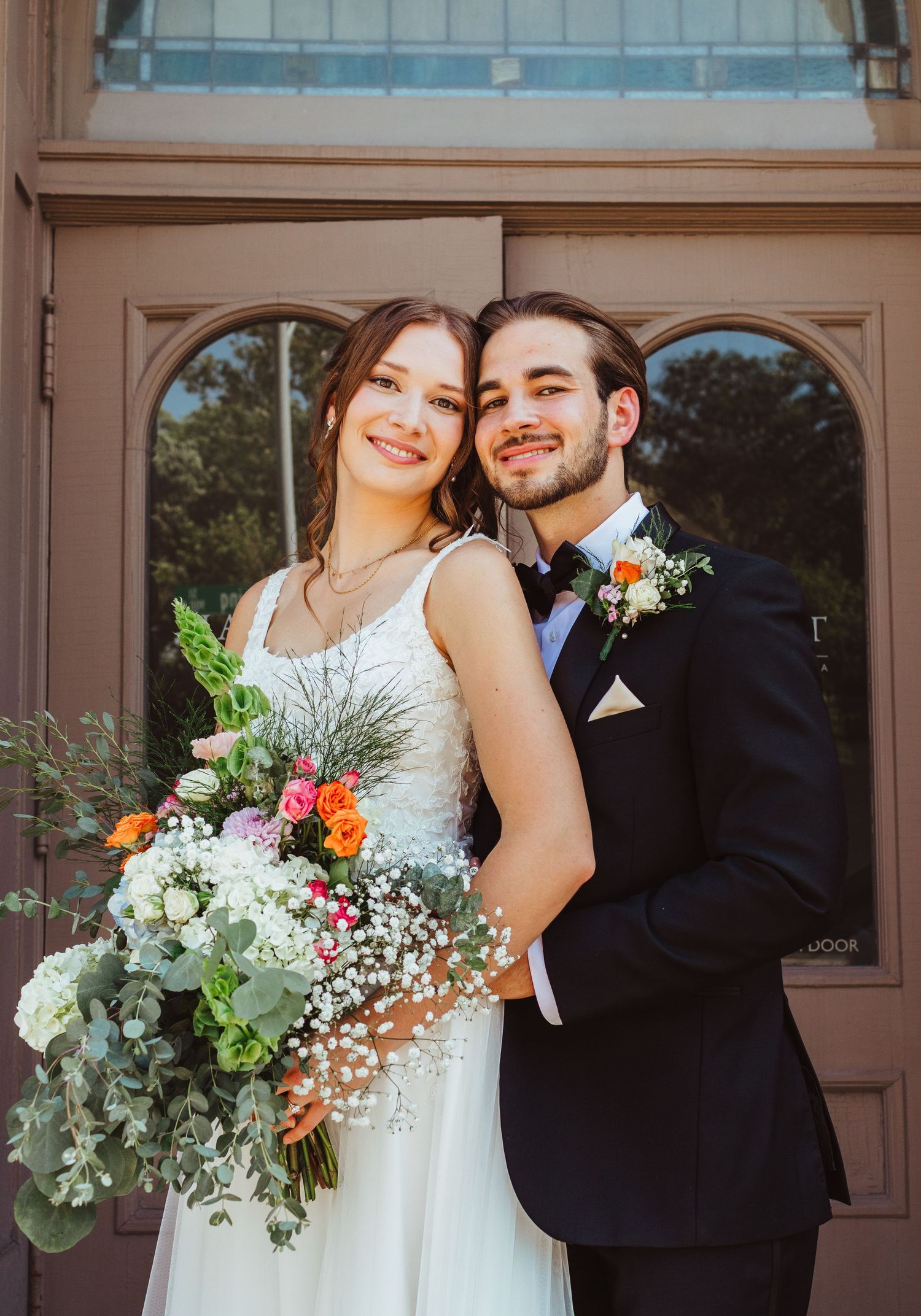 Smiling bride and groom pose together outside church doors with colorful wildflower bouquet.