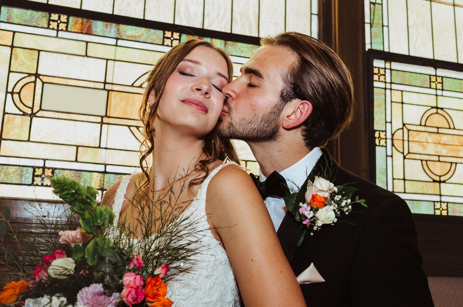 Groom tenderly kisses bride on cheek before colorful stained glass window inside church.