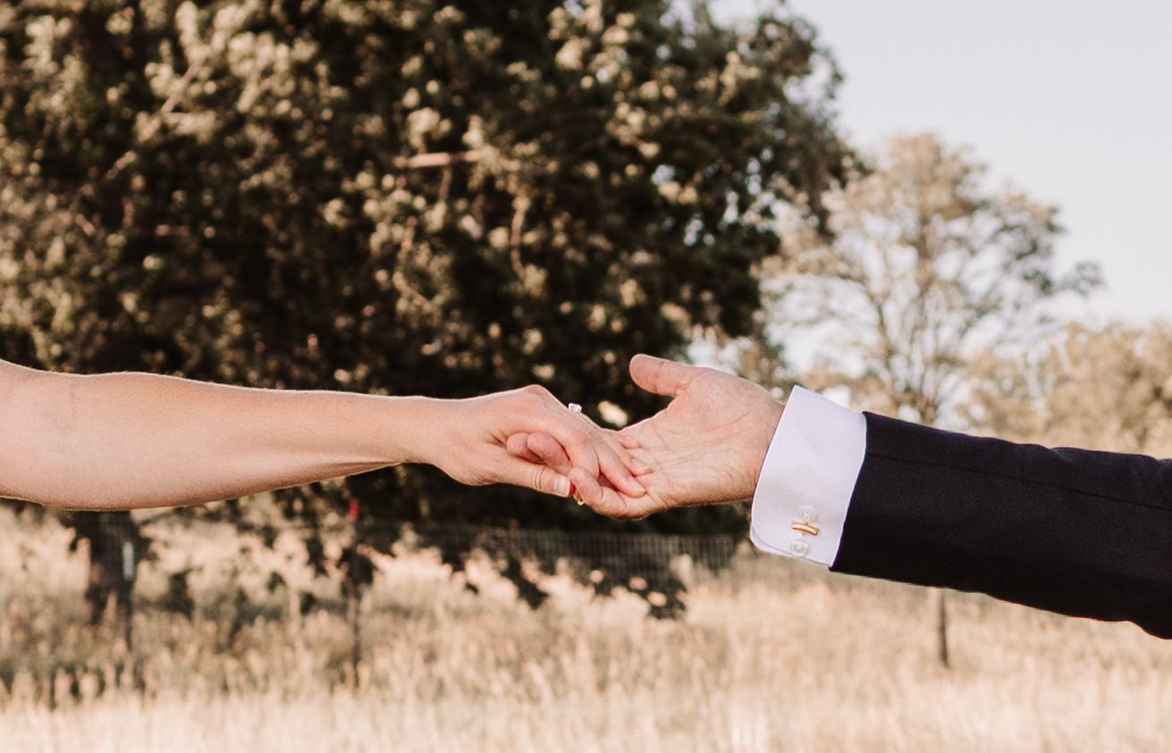 Bride and groom gently holding hands outdoors with golden trees in the background at a romantic wedding.