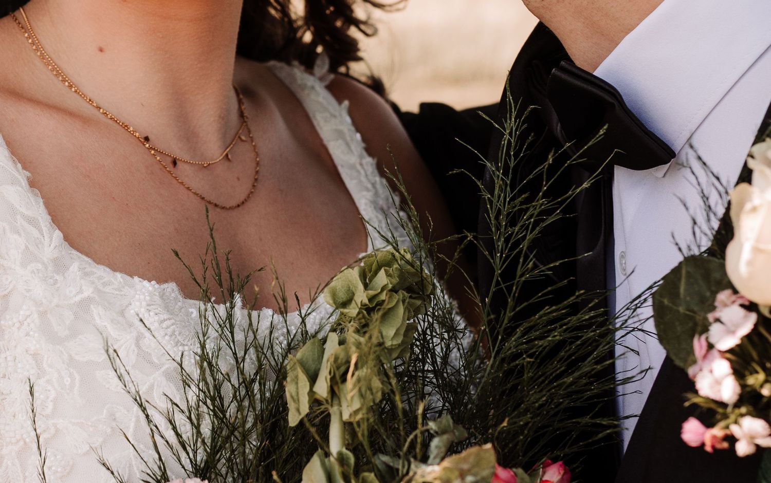 Close-up of bride in white lace gown wearing layered gold necklaces holding a lush wedding bouquet.