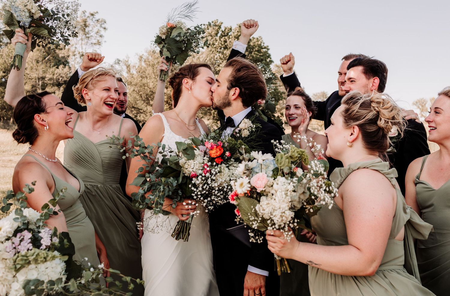 Joyful wedding party celebrating as bride and groom kiss surrounded by cheering bridesmaids in sage green dresses.