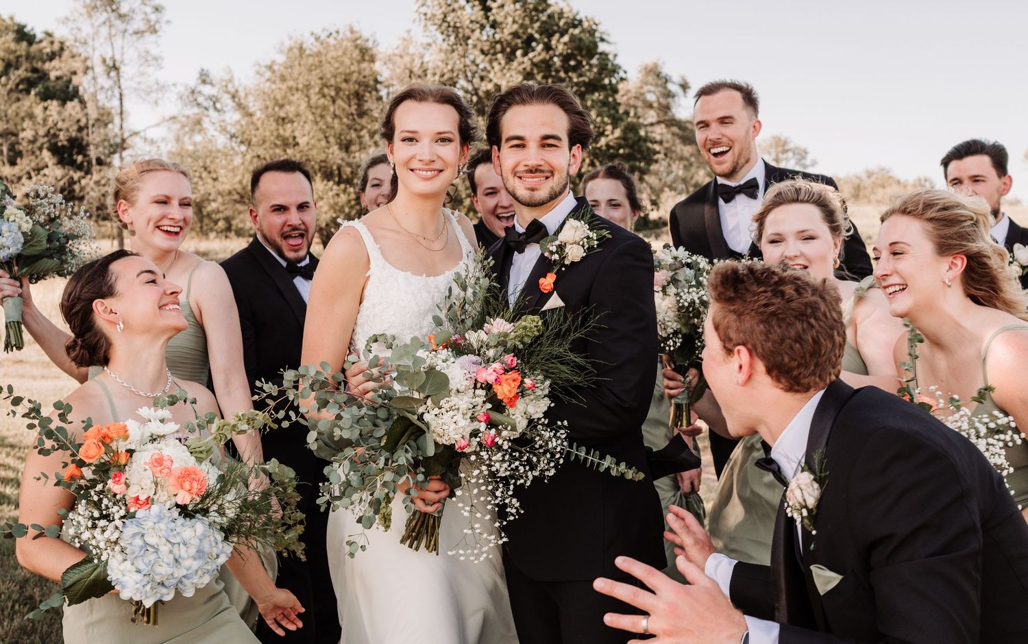 Smiling bride and groom pose with their laughing wedding party outdoors holding colorful floral bouquets.