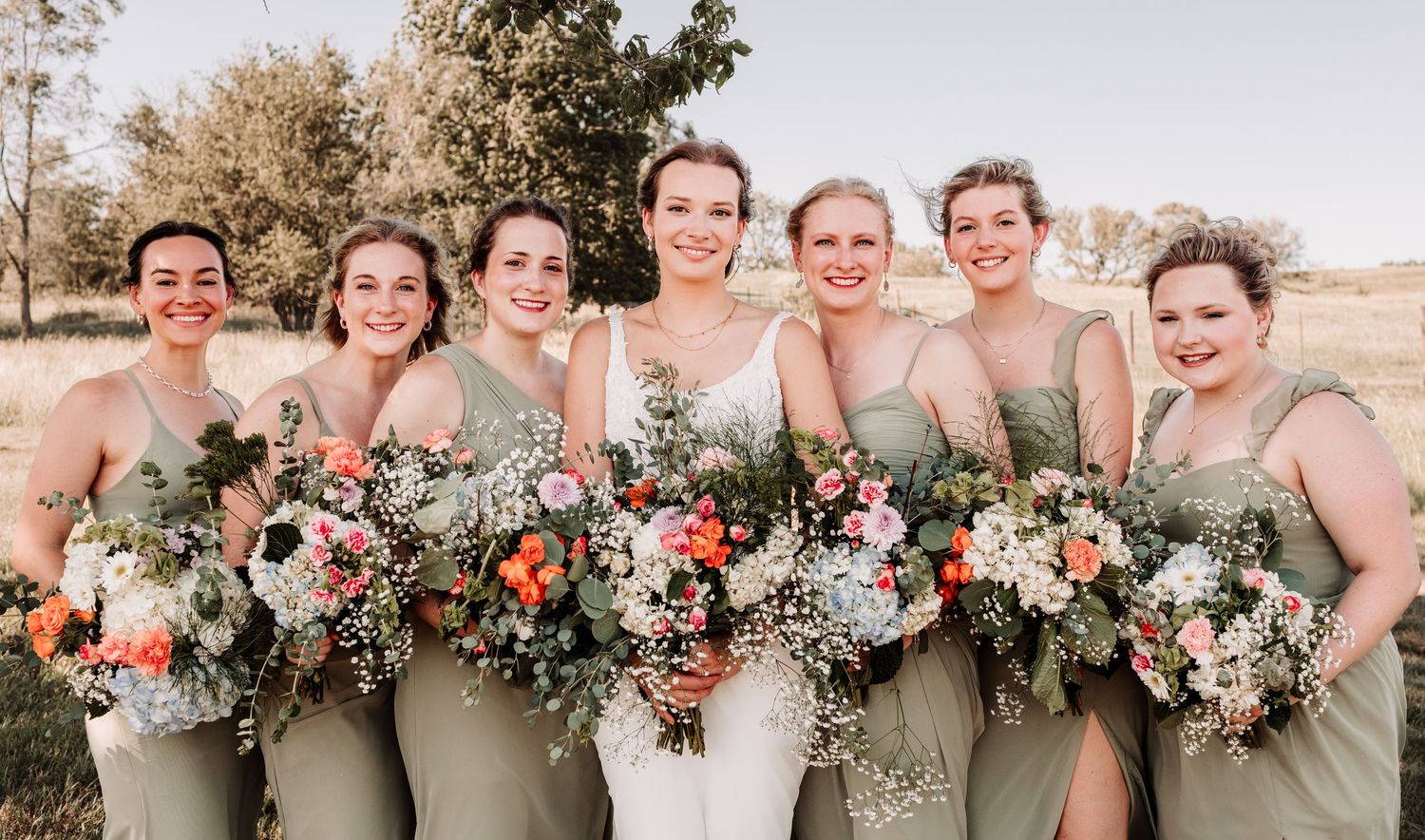 Bride stands with six bridesmaids in matching sage green dresses holding vibrant colorful floral bouquets outdoors.