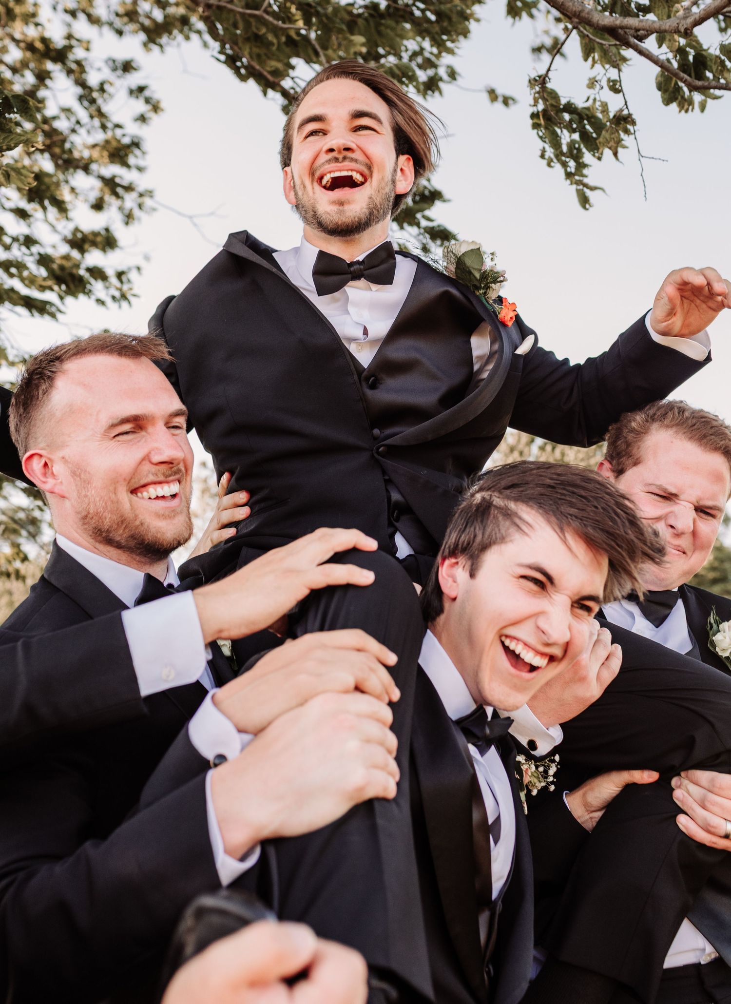 Groomsmen in black tuxedos joyfully lifting the groom on their shoulders in a celebratory outdoor photo.