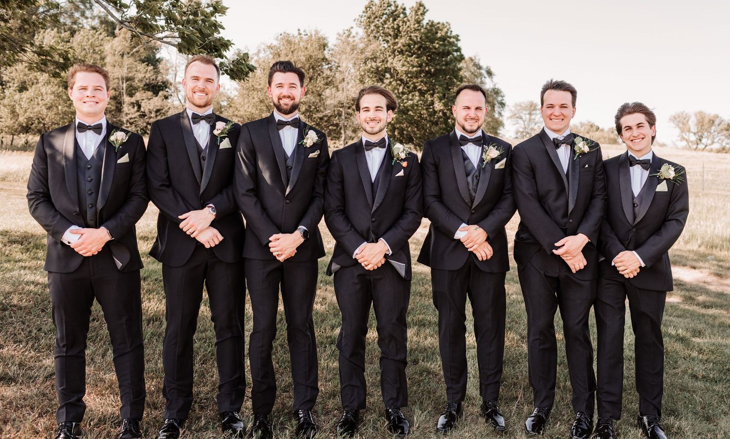 Seven groomsmen in matching black tuxedos with bow ties and boutonnieres posing outdoors on a green lawn.