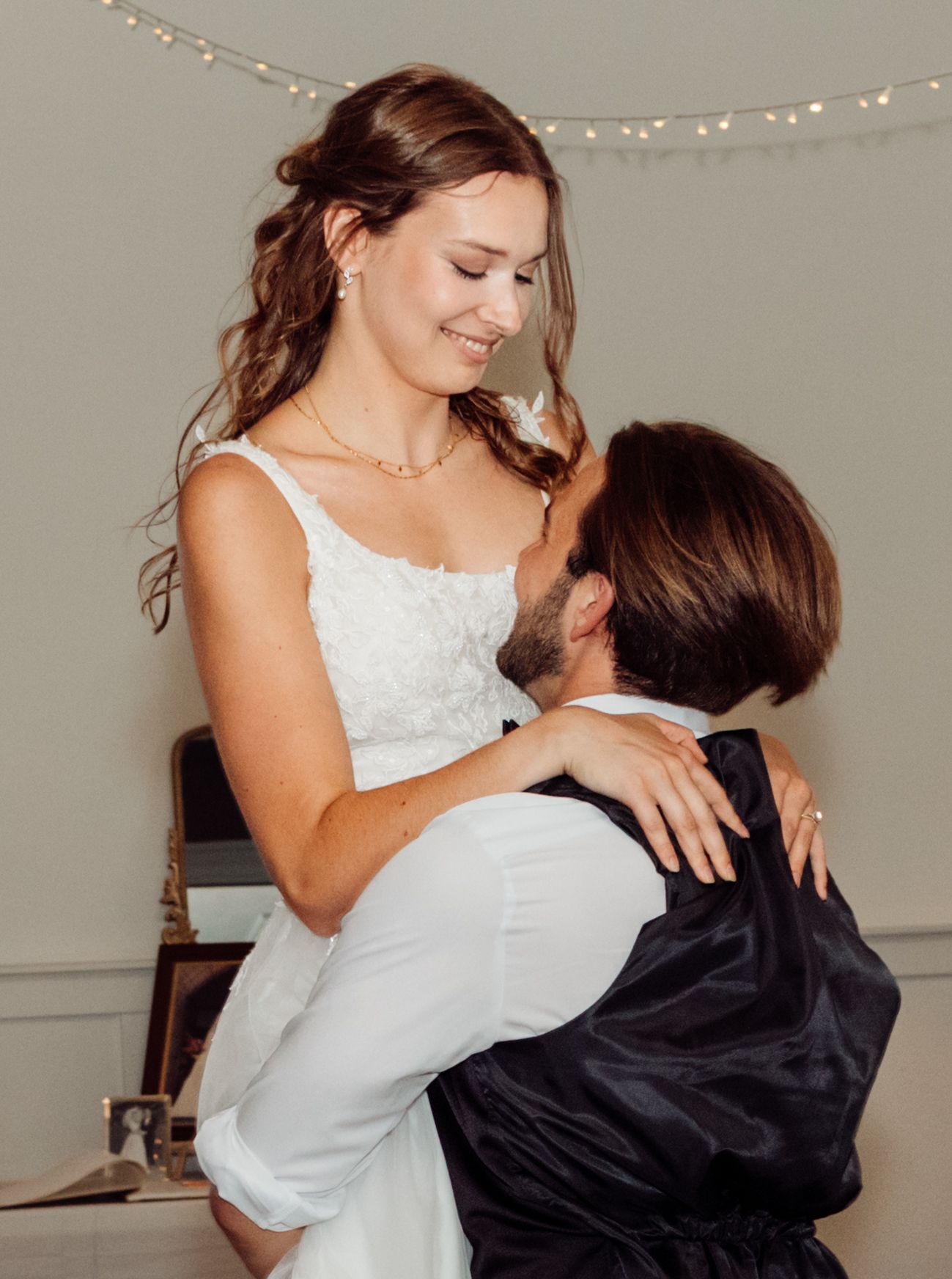 Groom lifts smiling bride in a white wedding dress under warm string lights during a romantic indoor moment.
