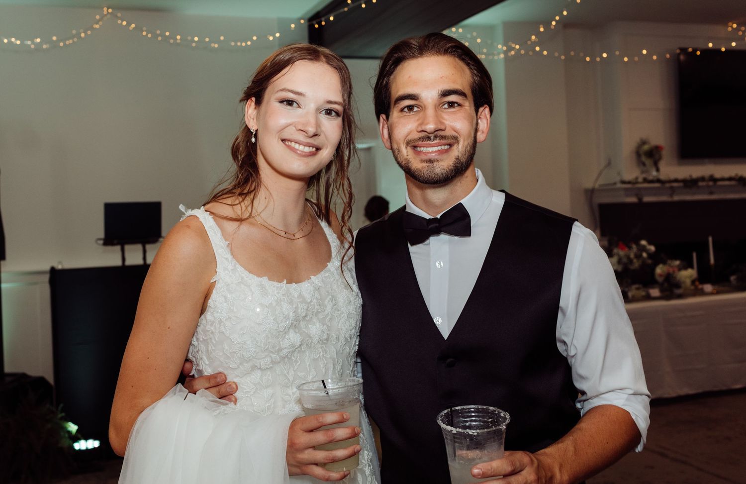 Bride and groom smile together holding drinks at their wedding reception under twinkling string lights indoors.
