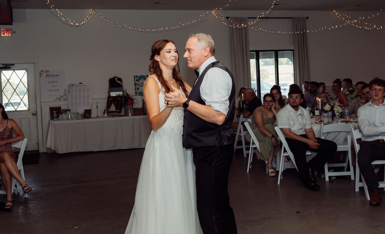 Father and daughter share an emotional first dance at a wedding reception with guests seated in background.