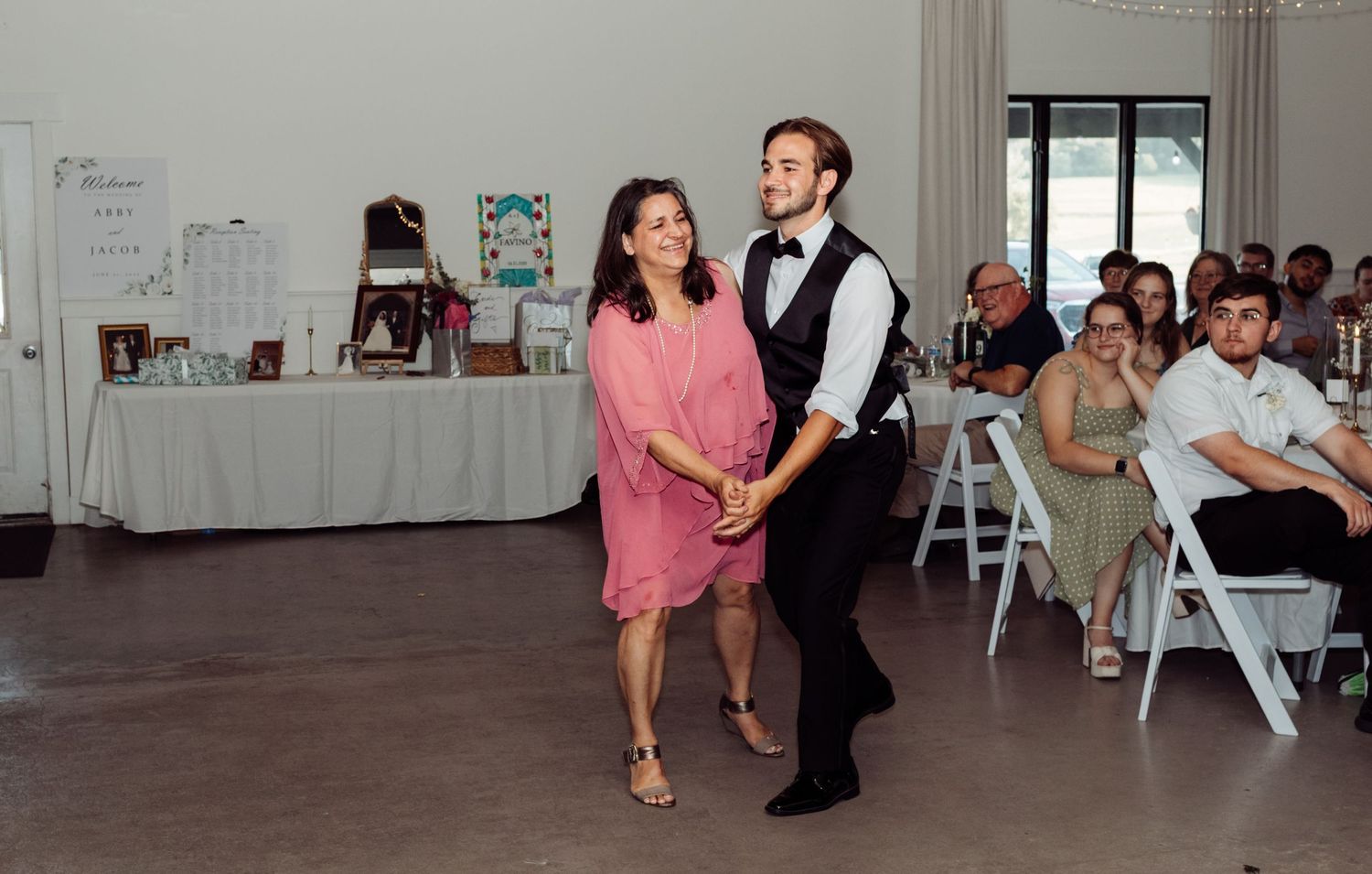 A young man in a vest dances with a woman in a pink dress at a wedding reception.