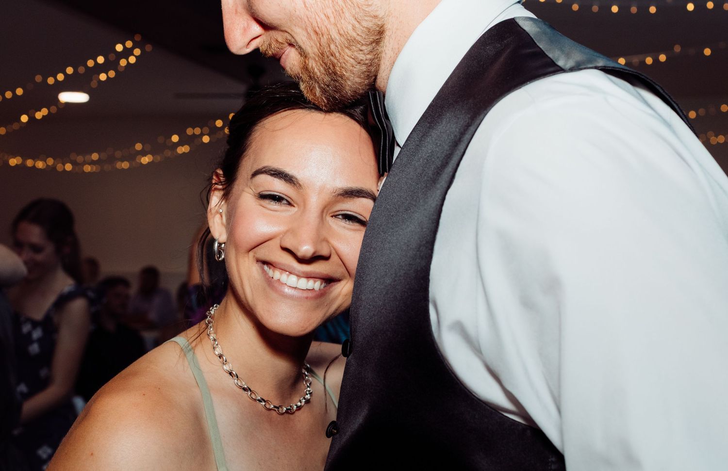 A smiling bride in a sage dress dances closely with a groom in a black vest at a wedding.