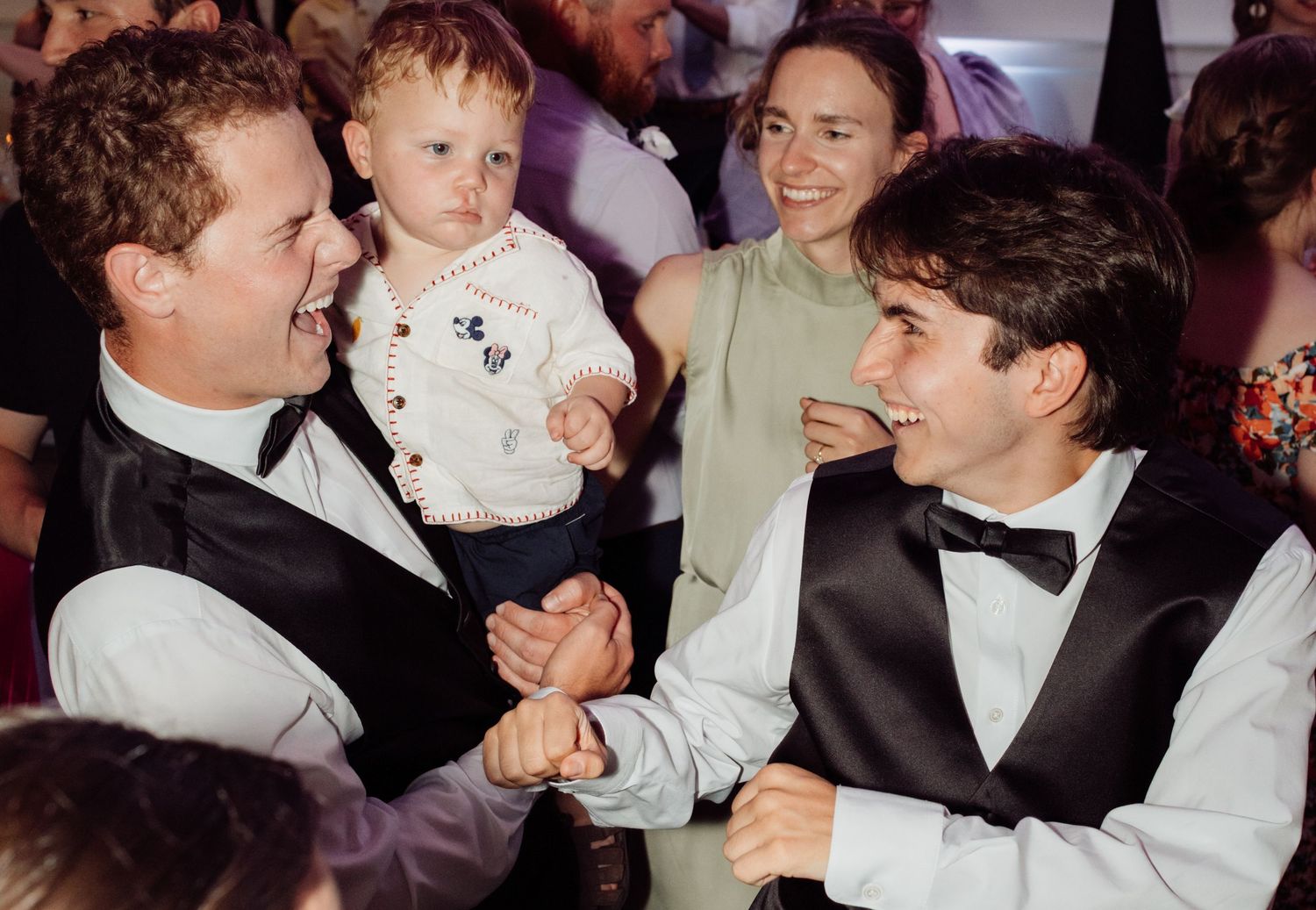 Two men in black vests shake hands and laugh at a wedding reception, one holding a toddler.