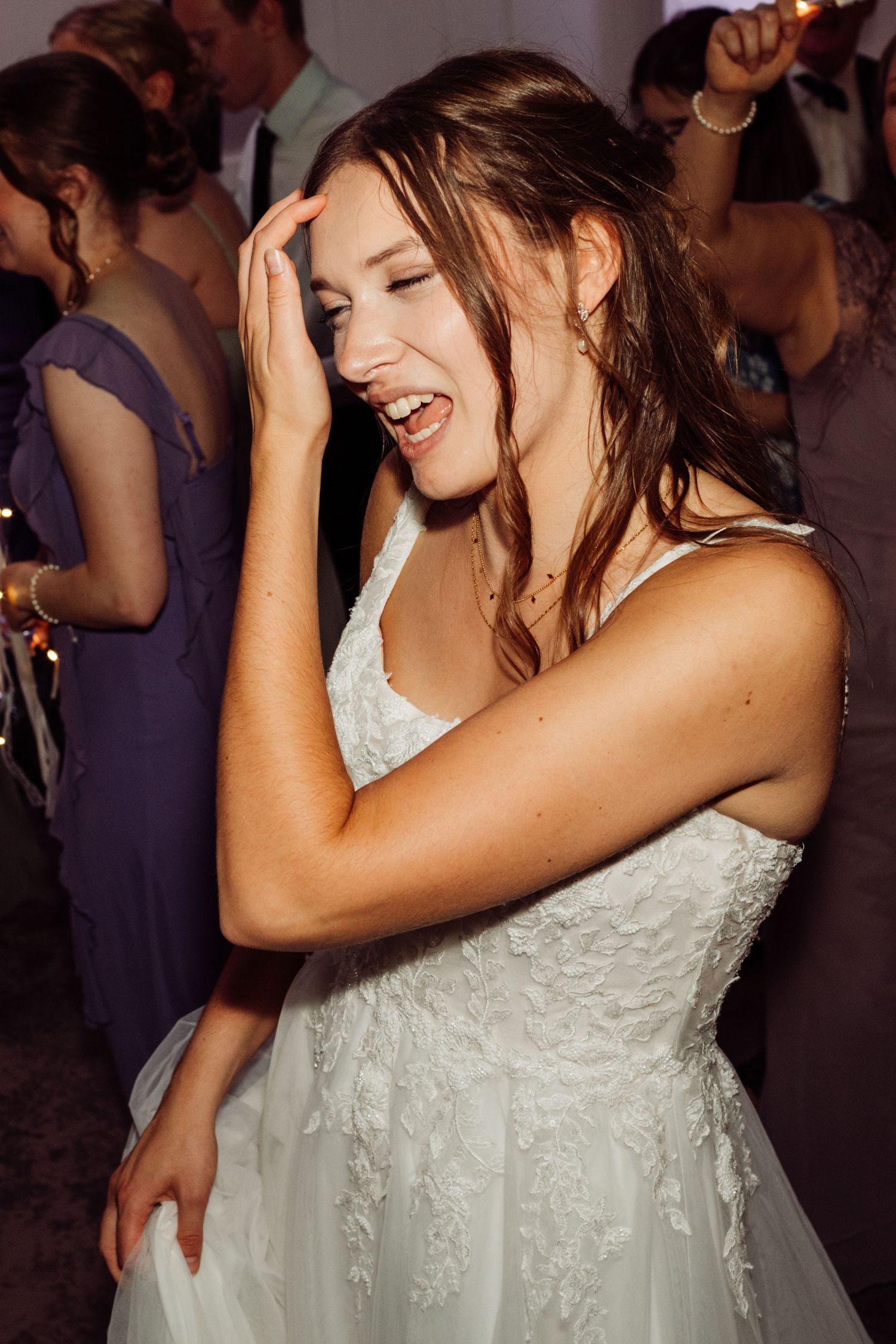 A bride in a white lace gown laughs joyfully while dancing at her wedding reception.