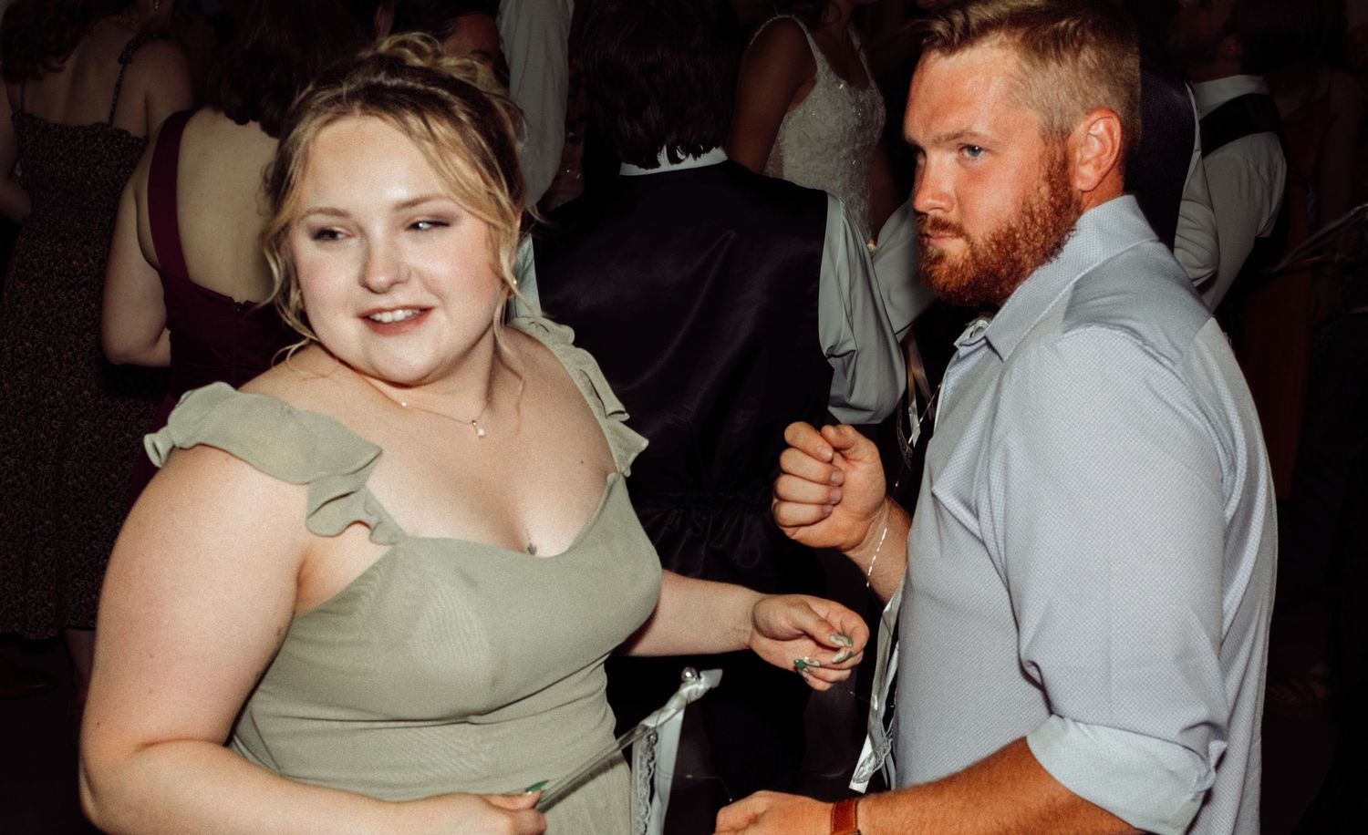 A woman in a sage off-shoulder dress dances with a bearded man in a gray shirt at a wedding.