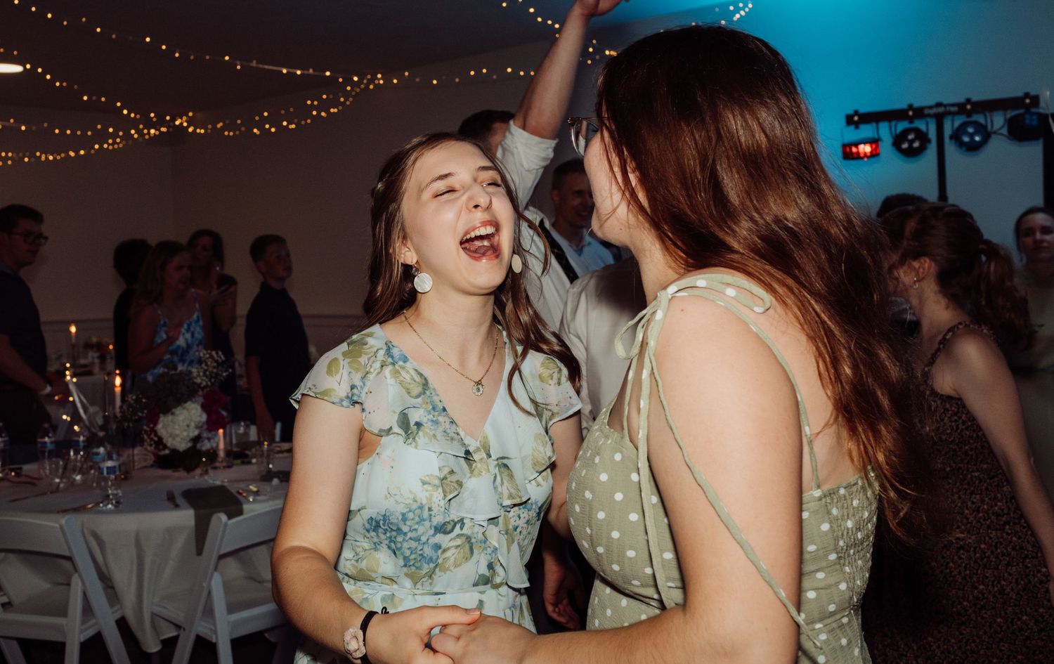 Two women in floral dresses laugh and dance together joyfully at a wedding reception.