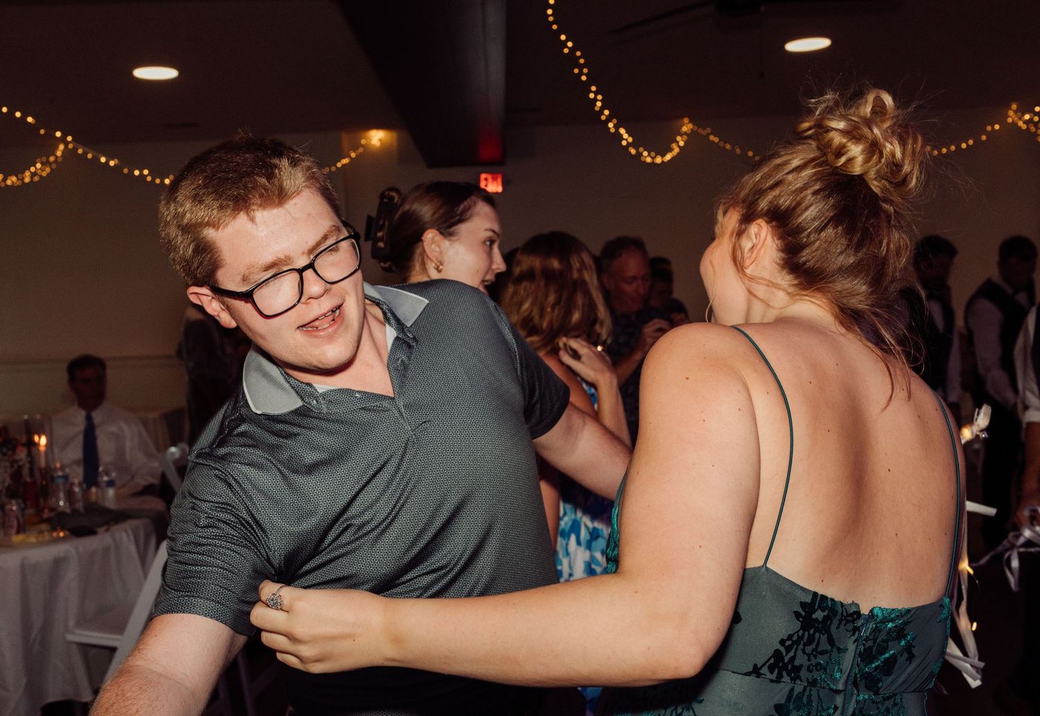 A young man in glasses dances with a woman in a green dress at a lively wedding reception.