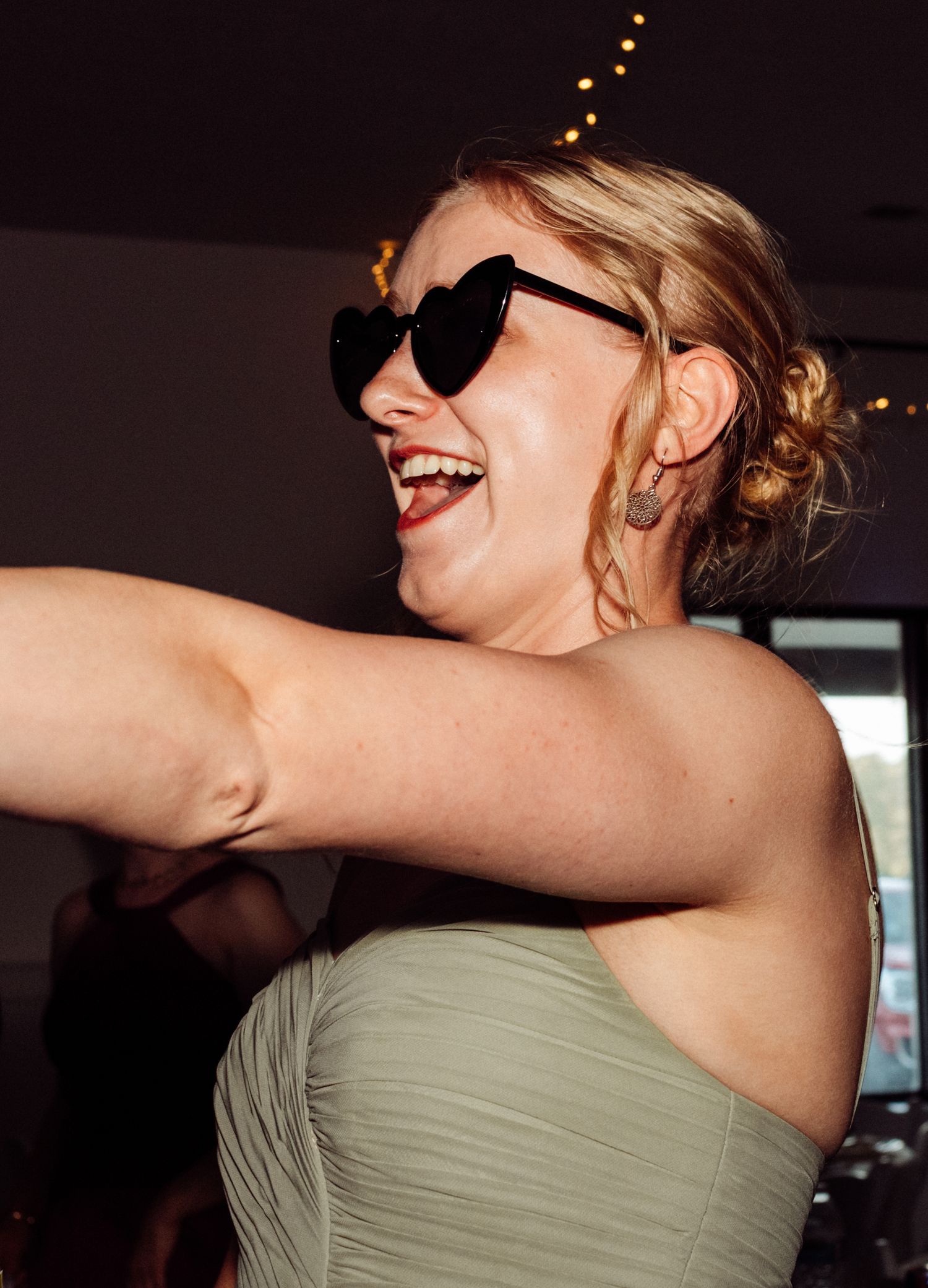 A blonde woman in a sage strapless dress laughs while wearing heart-shaped sunglasses at a wedding.