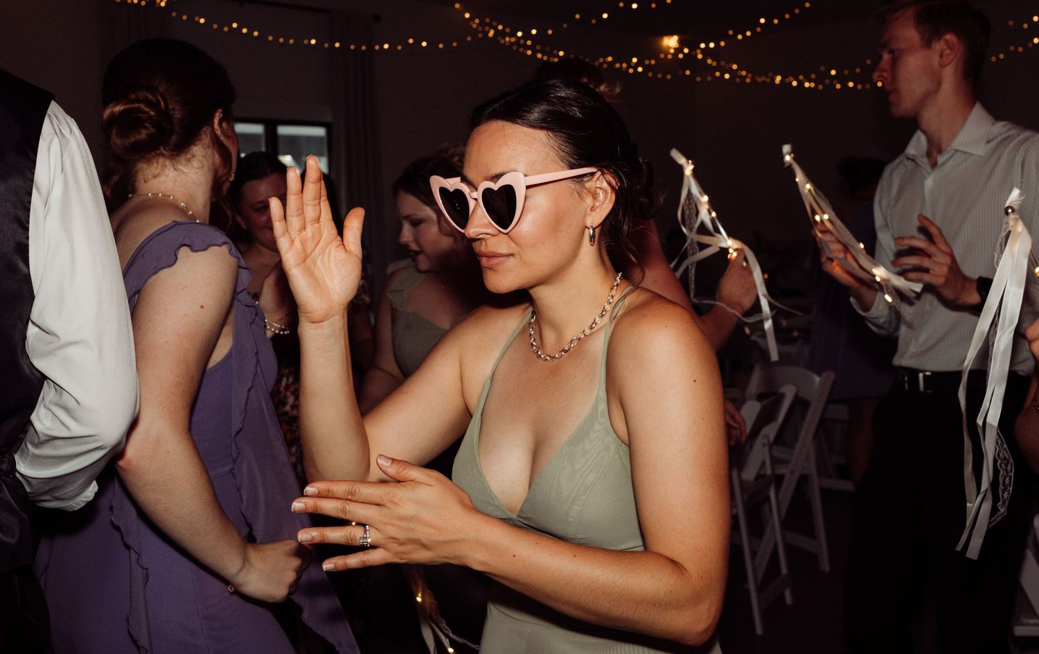 A woman in a sage dress dances wearing heart-shaped white sunglasses at a wedding reception.