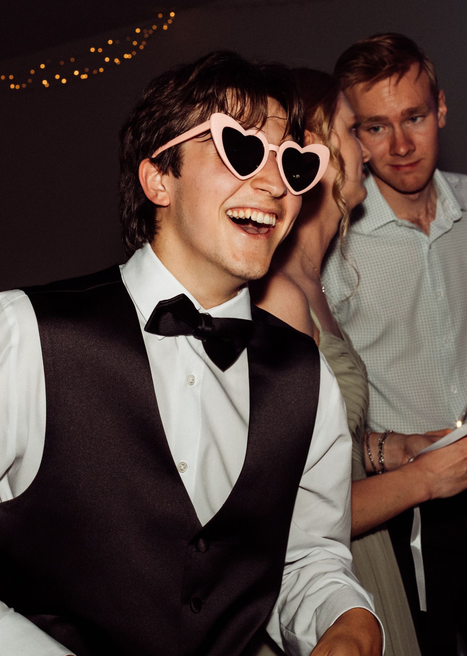 Laughing young man wearing pink heart-shaped sunglasses and a black bow tie vest at a wedding reception.