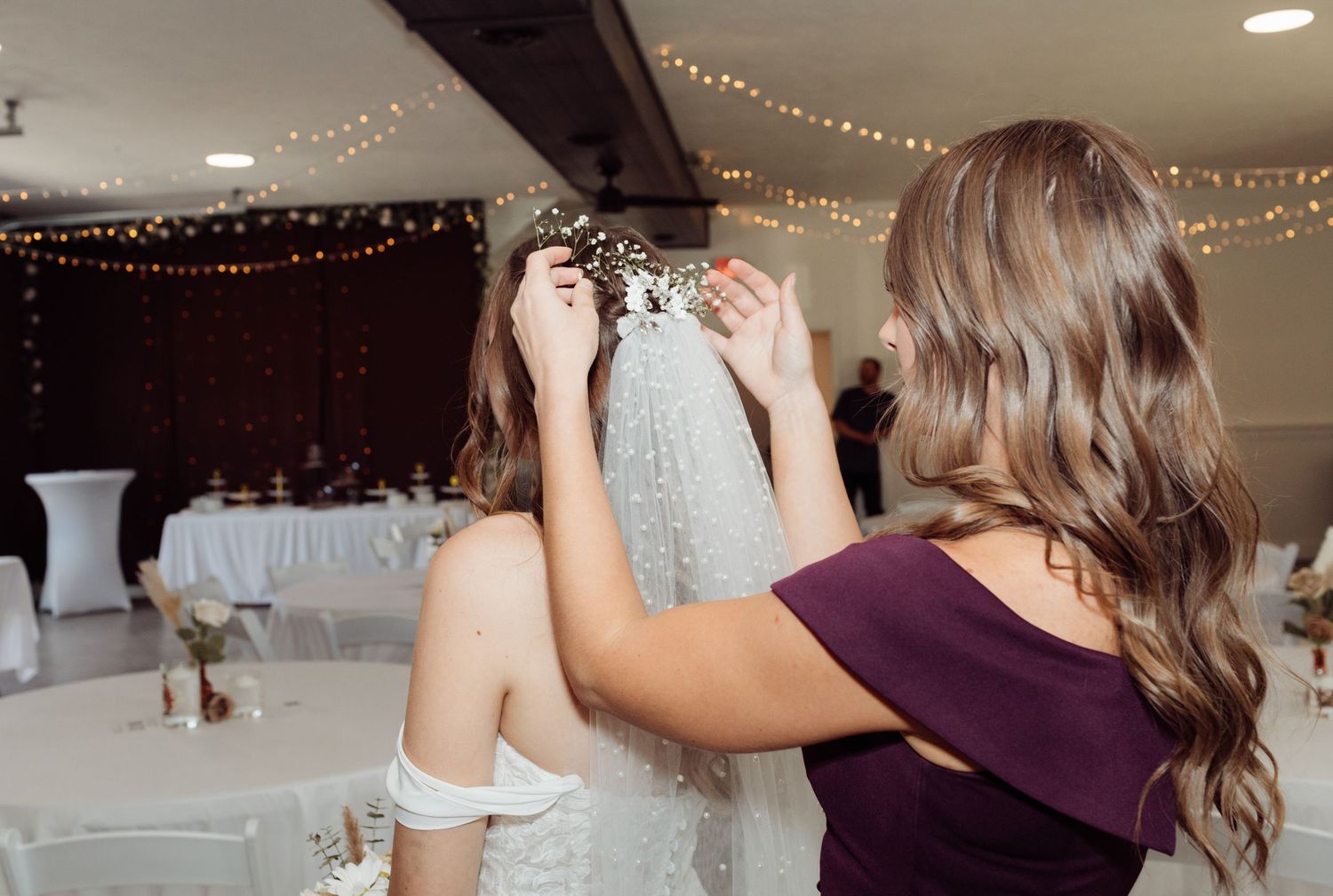 Bridesmaid carefully adjusting the veil of a bride in an elegantly decorated wedding reception hall.