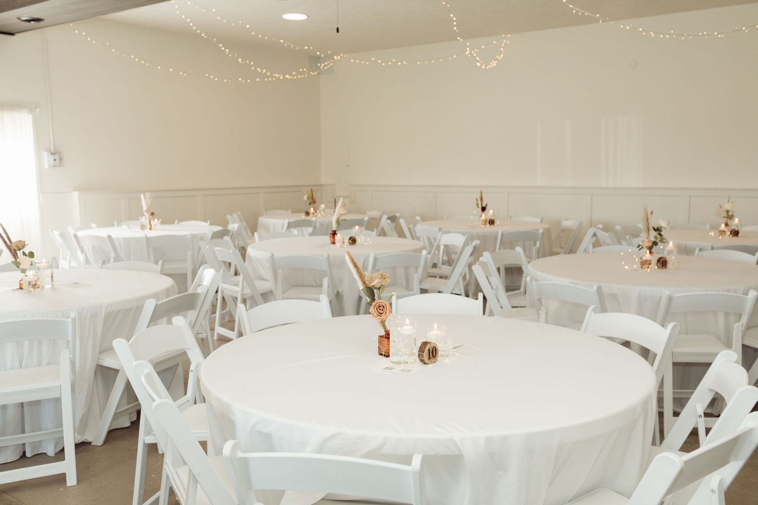 Elegant wedding reception hall with white round tables, white folding chairs, and soft string lights creating a romantic atmosphere.