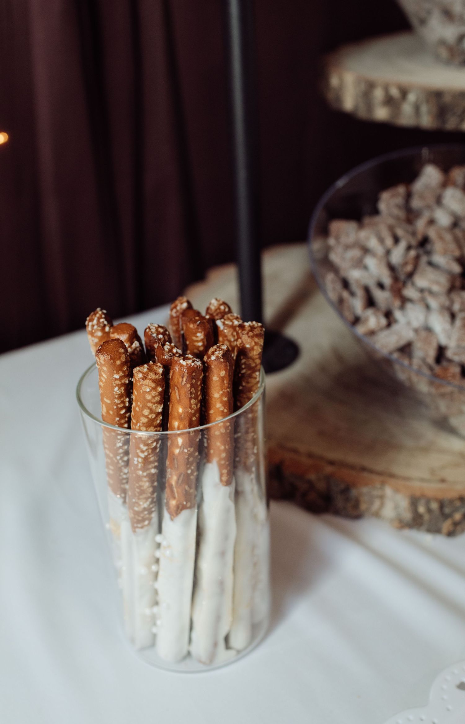 White chocolate-dipped pretzel rods standing upright in a clear glass vase on a white tablecloth at a wedding reception.